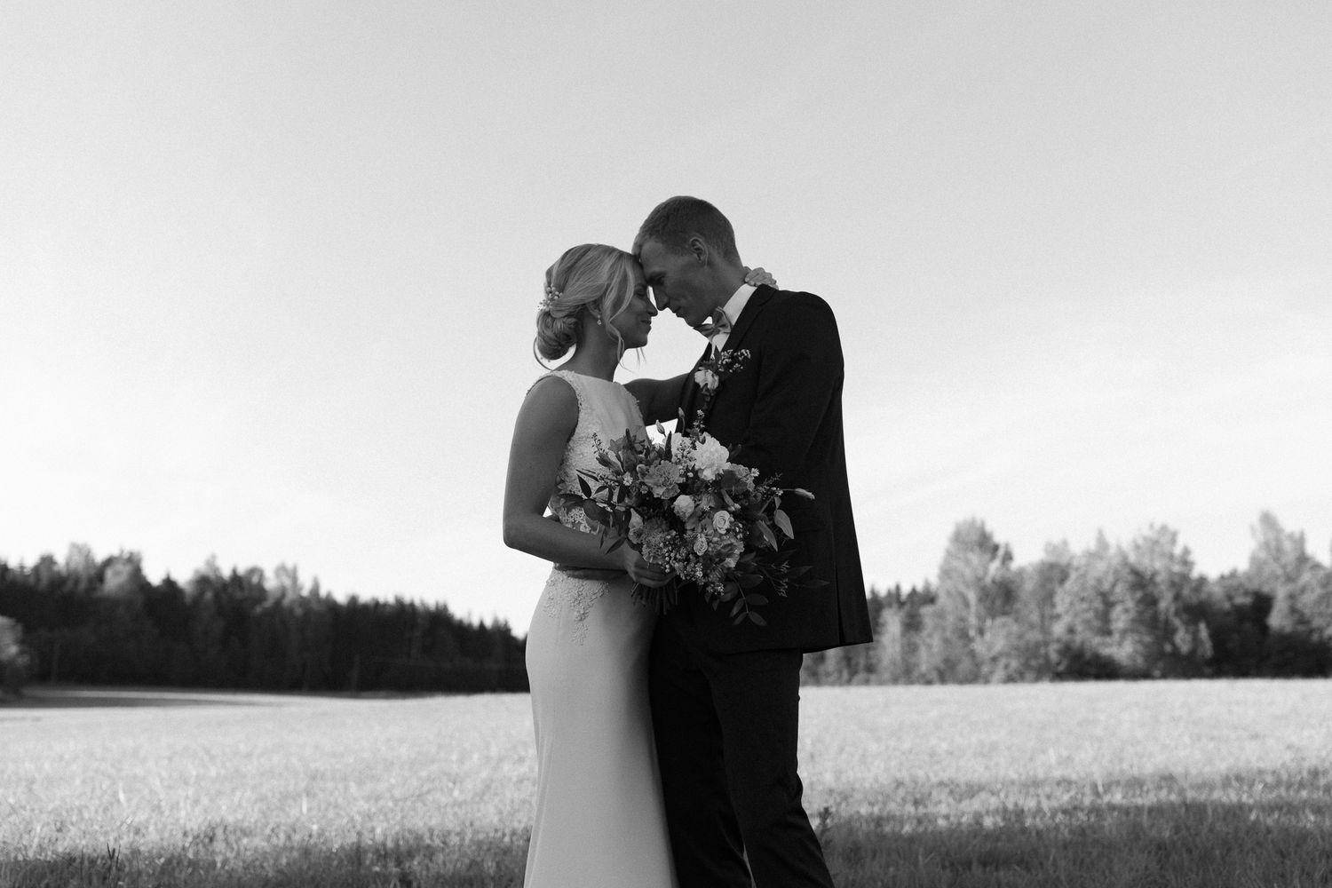 Romantic black and white portrait of newlyweds embracing in a meadow with forest backdrop.
