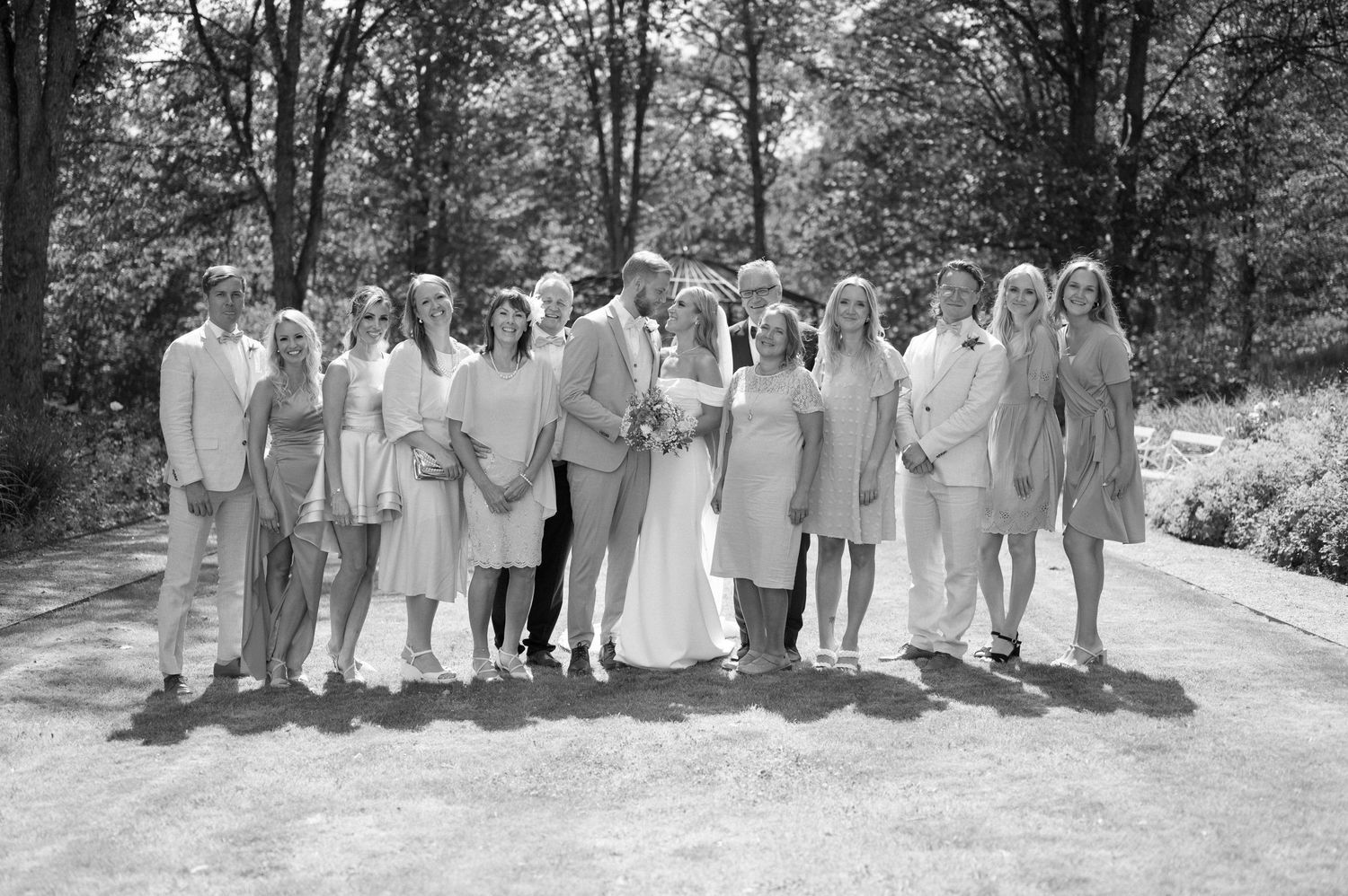 Wedding party in light colored attire poses outdoors on a sunny day for group photos.