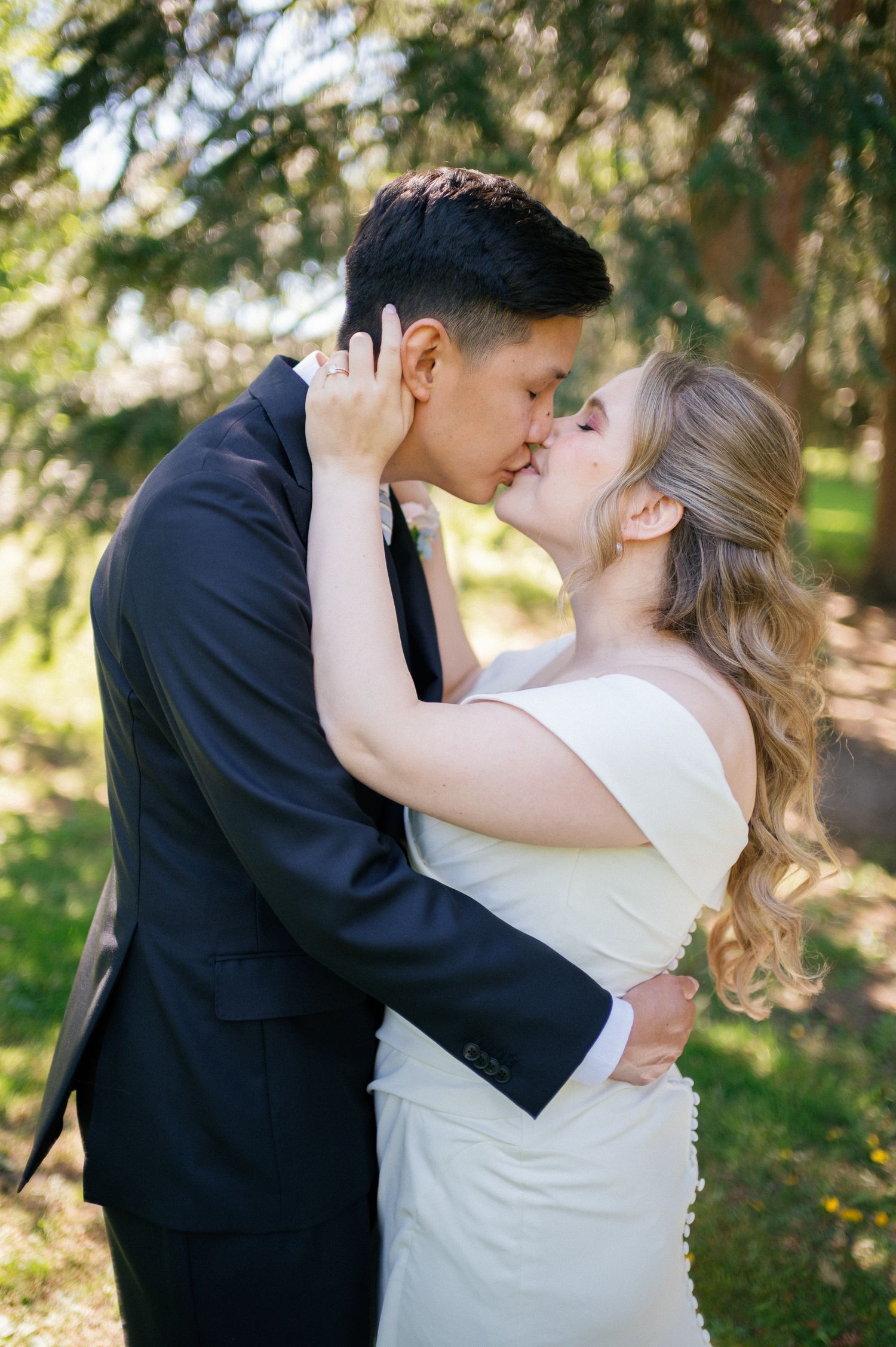 A bride and groom share a romantic first kiss outdoors surrounded by lush green trees on their wedding day.