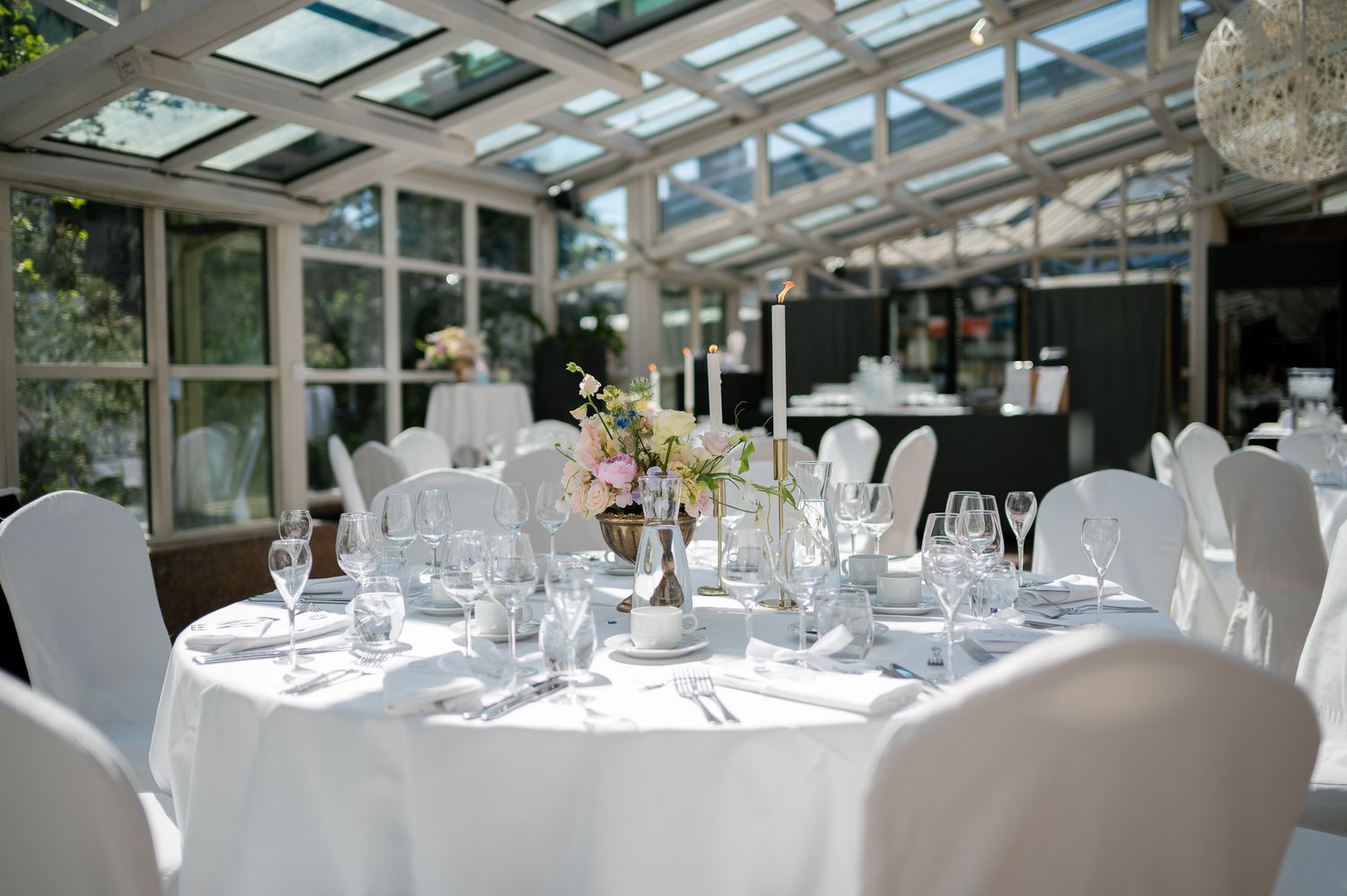 Elegant wedding reception in a sunlit conservatory with white round tables, floral centerpieces, and white chair covers.