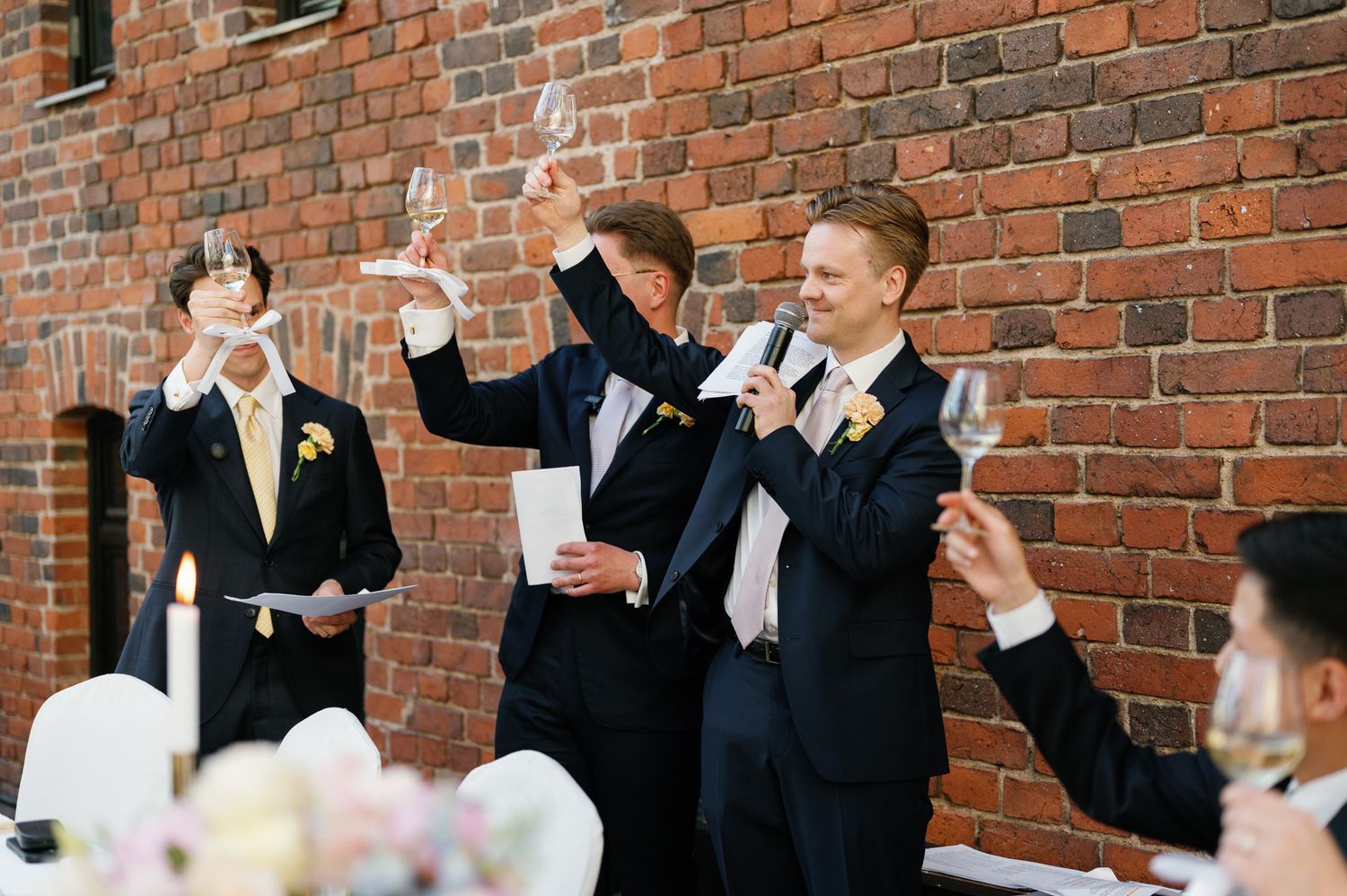 Men in tuxedos raising champagne glasses during a wedding toast at a brick wall venue.