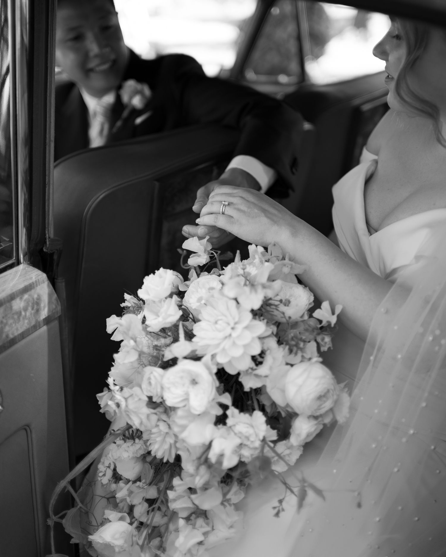 Black and white photo of a bride holding a lush floral bouquet beside a groom in a vintage car.