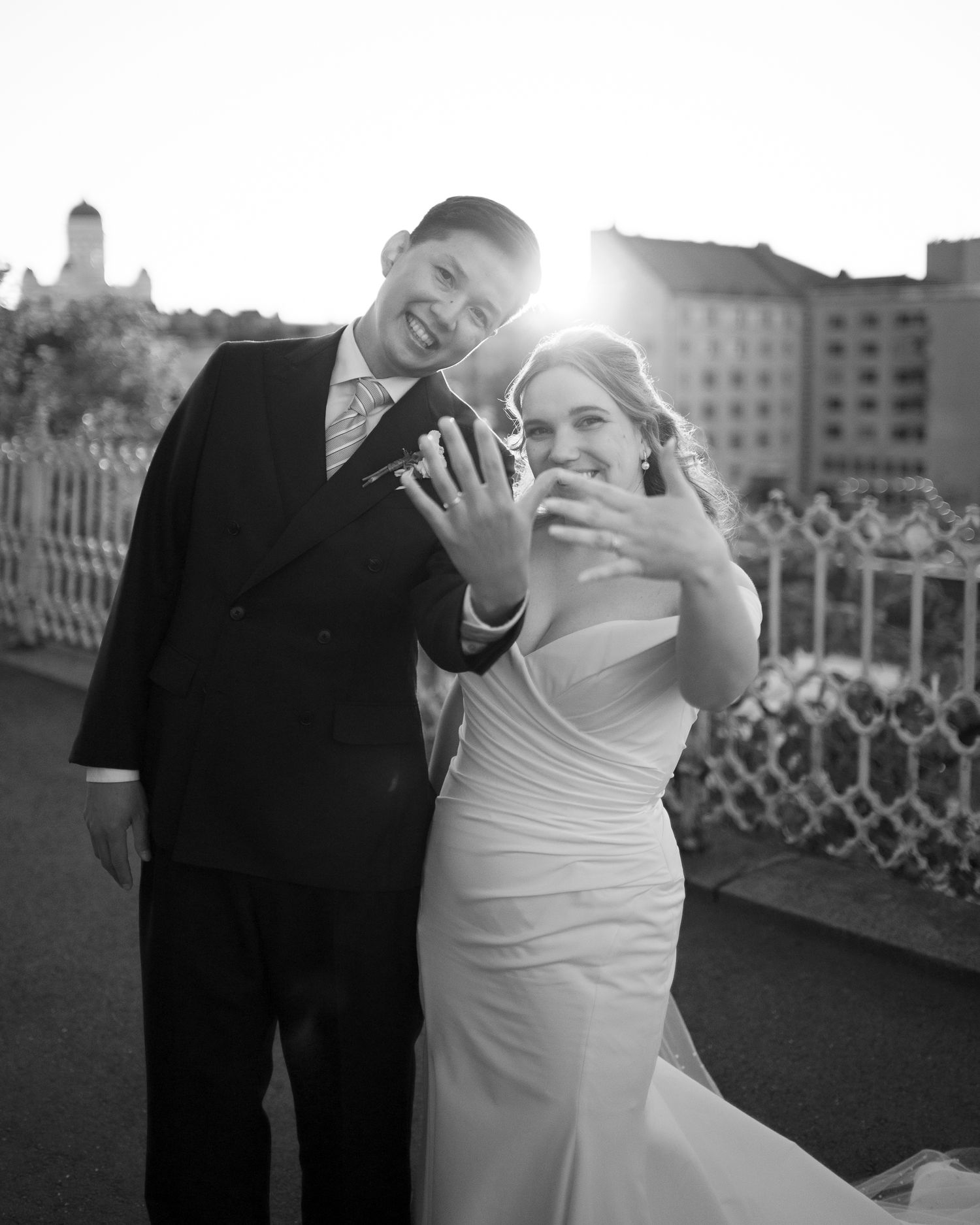 Black and white photo of newlyweds showing off wedding rings, smiling outdoors with sunburst and buildings behind them.