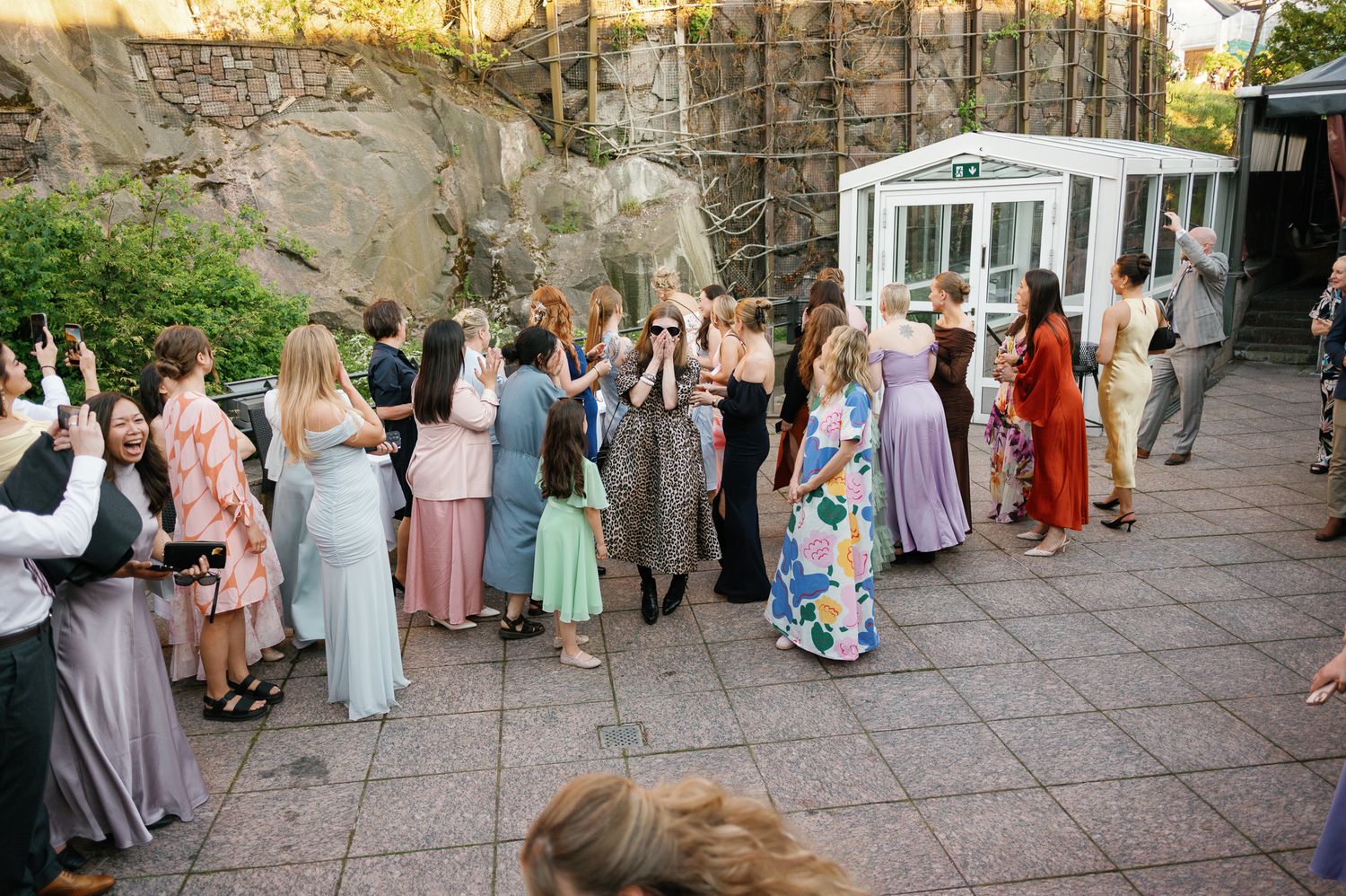 Wedding guests in colorful formal attire gather outdoors on a patio, socializing and celebrating near a white building.
