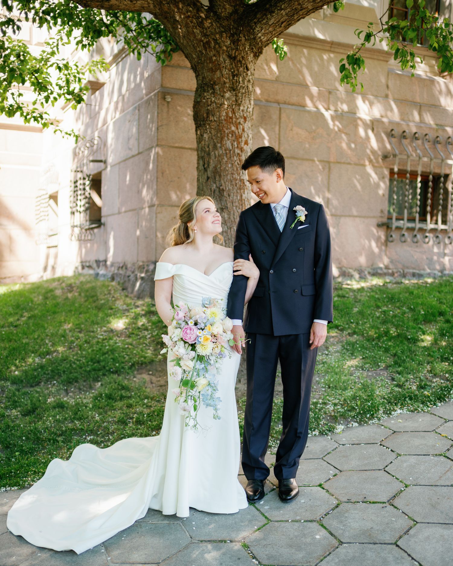 A bride in a white off-shoulder gown holds a pastel bouquet beside a groom in a navy suit under a tree.