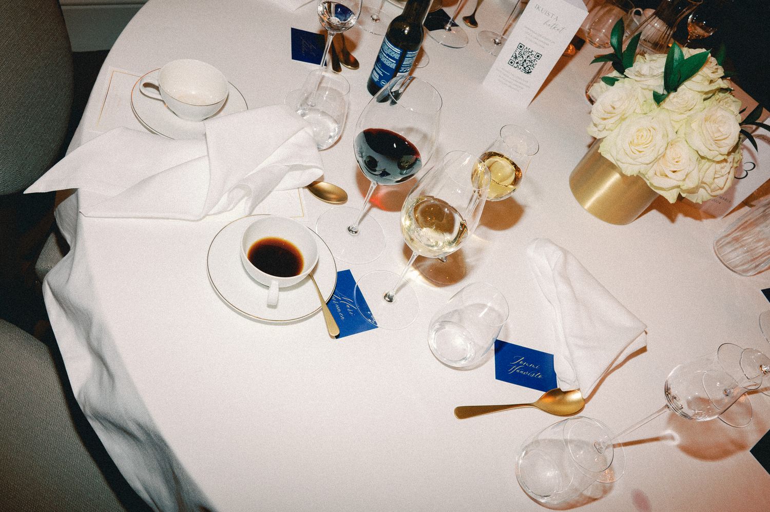 Wine glasses, coffee cups, and table settings arranged on a white tablecloth at a formal dining event.