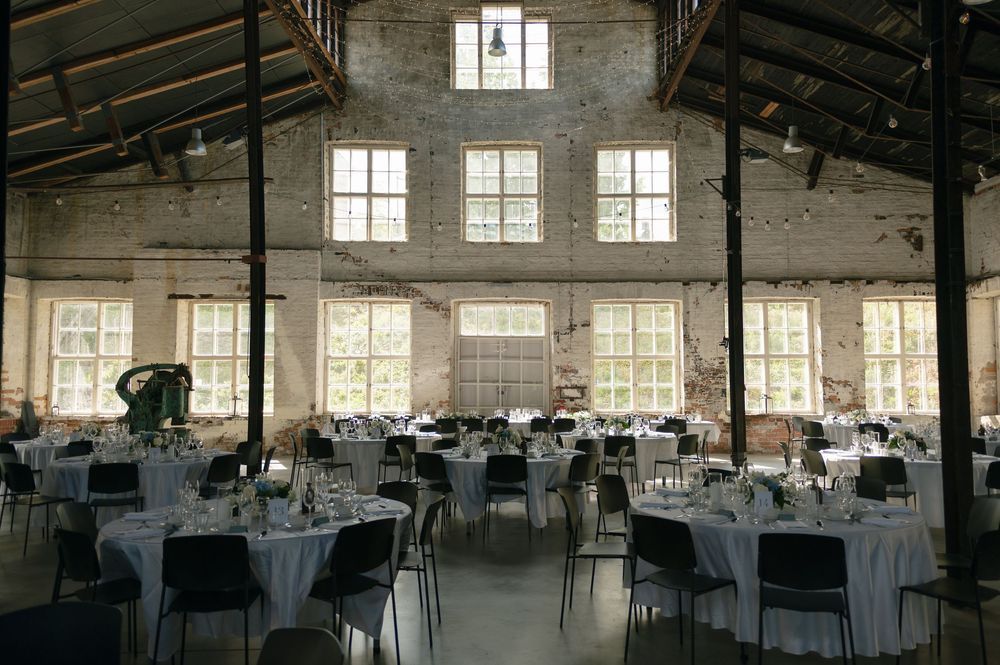 Spacious warehouse-style event space with white-clothed tables and natural lighting.