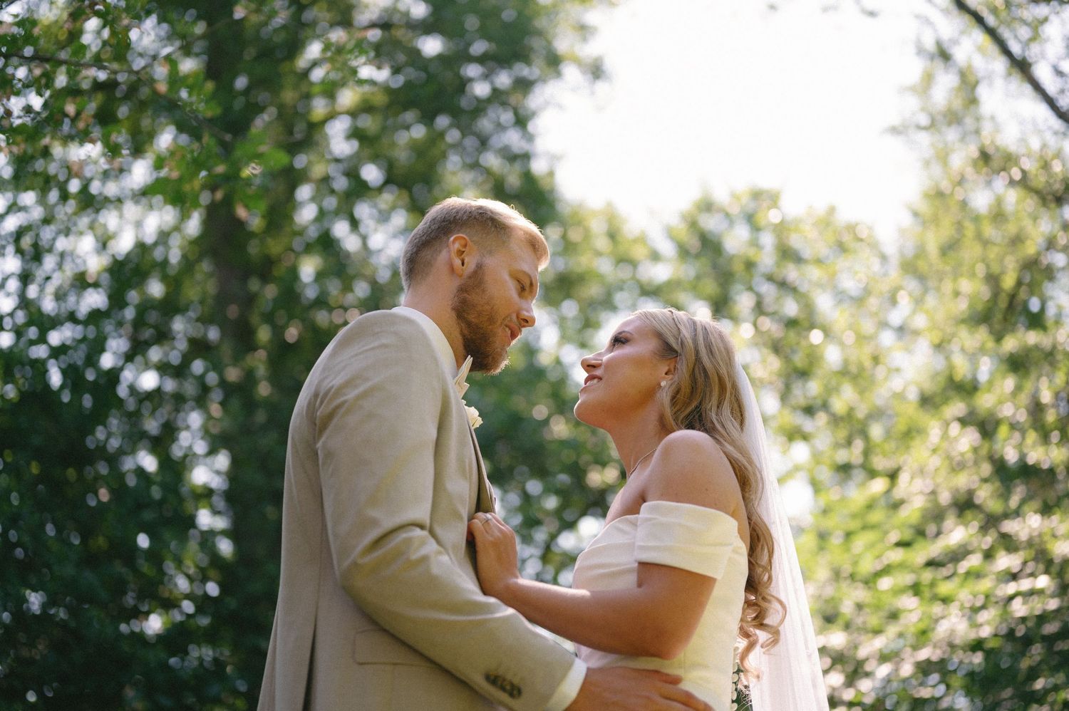 Couple embraces outdoors under sunlit trees during summer wedding.
