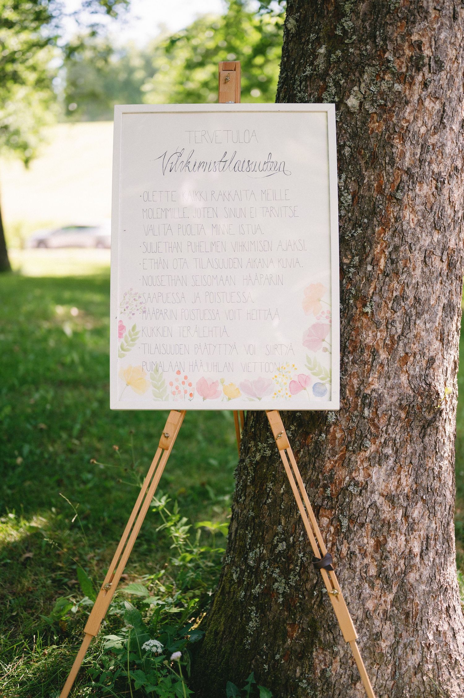 Wedding signage displayed on wooden easels against tree trunks in garden setting.