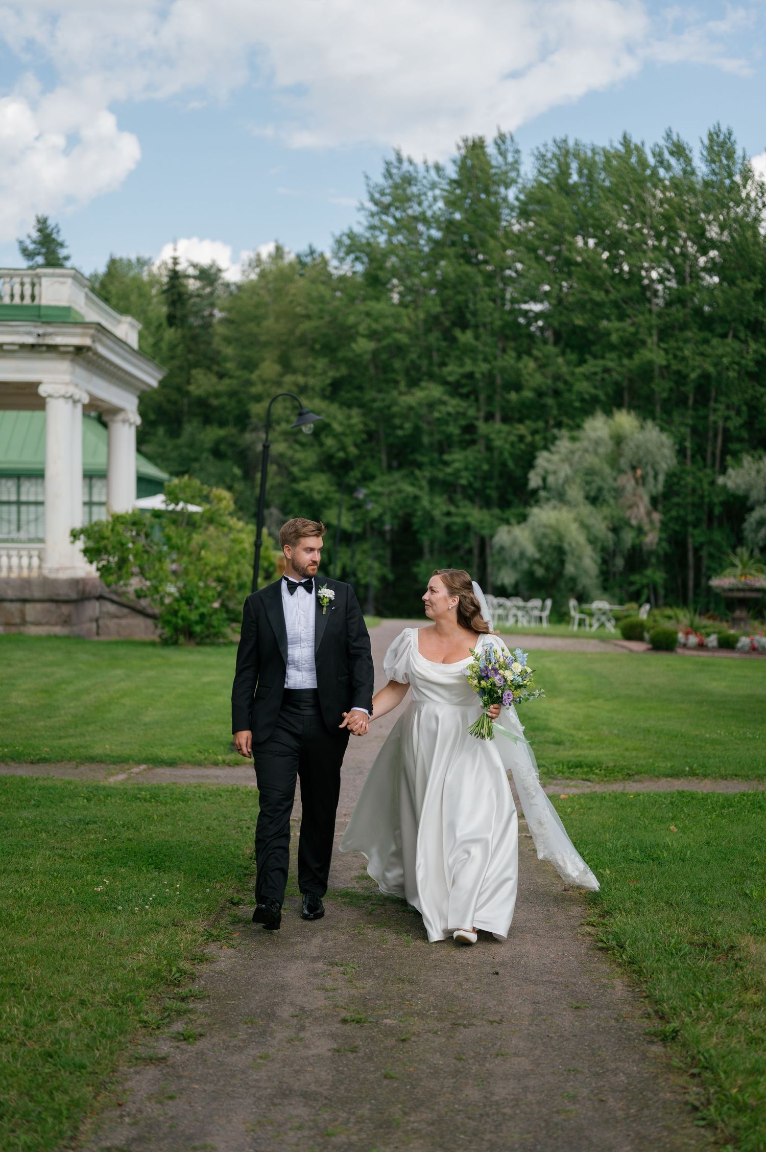 Wedding couple walking along a path with buildings and trees in the background.