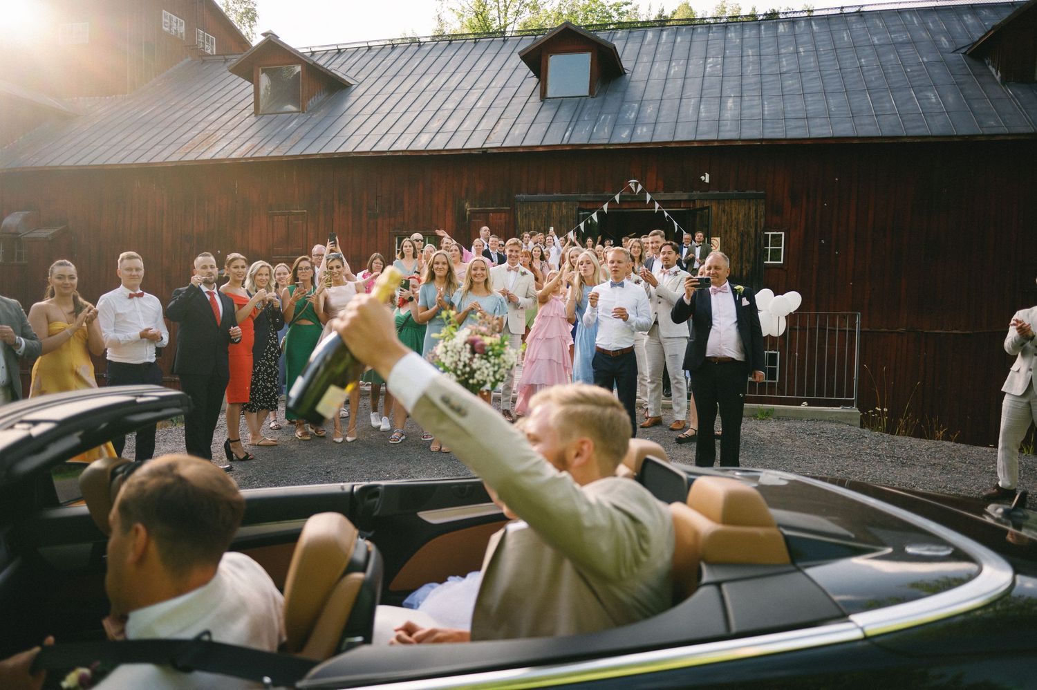 Wedding couple waves from convertible car as guests celebrate their departure.