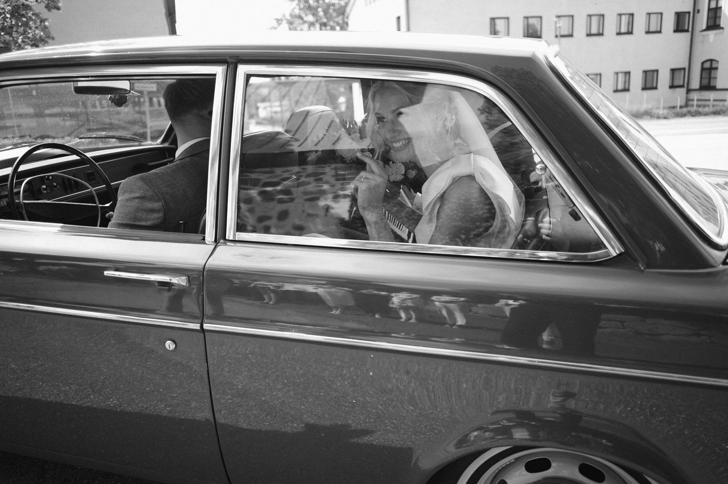A black and white photograph shows passengers seated in a vintage car looking through the side window.