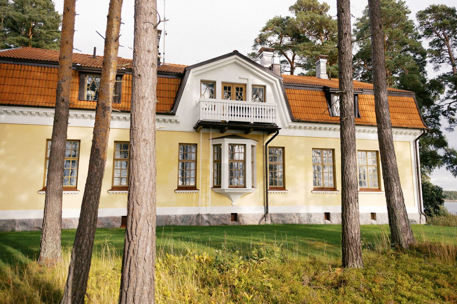 A panoramic view of a historic yellow building with orange roof tiles nestled among tall pine trees on a grassy hillside.