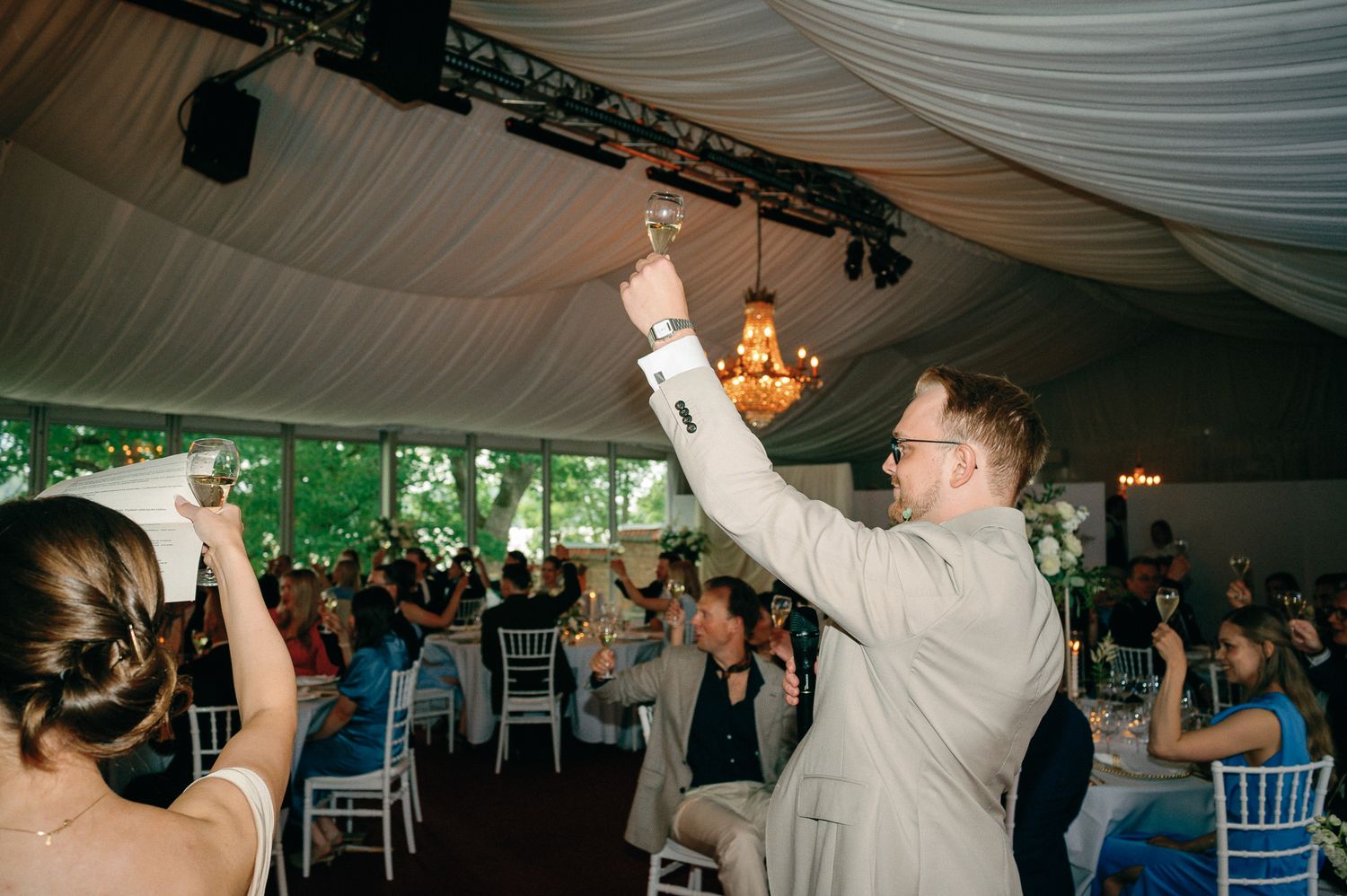 Bride and groom raising glasses in a toast at elegant tented wedding reception with chandelier and seated guests.