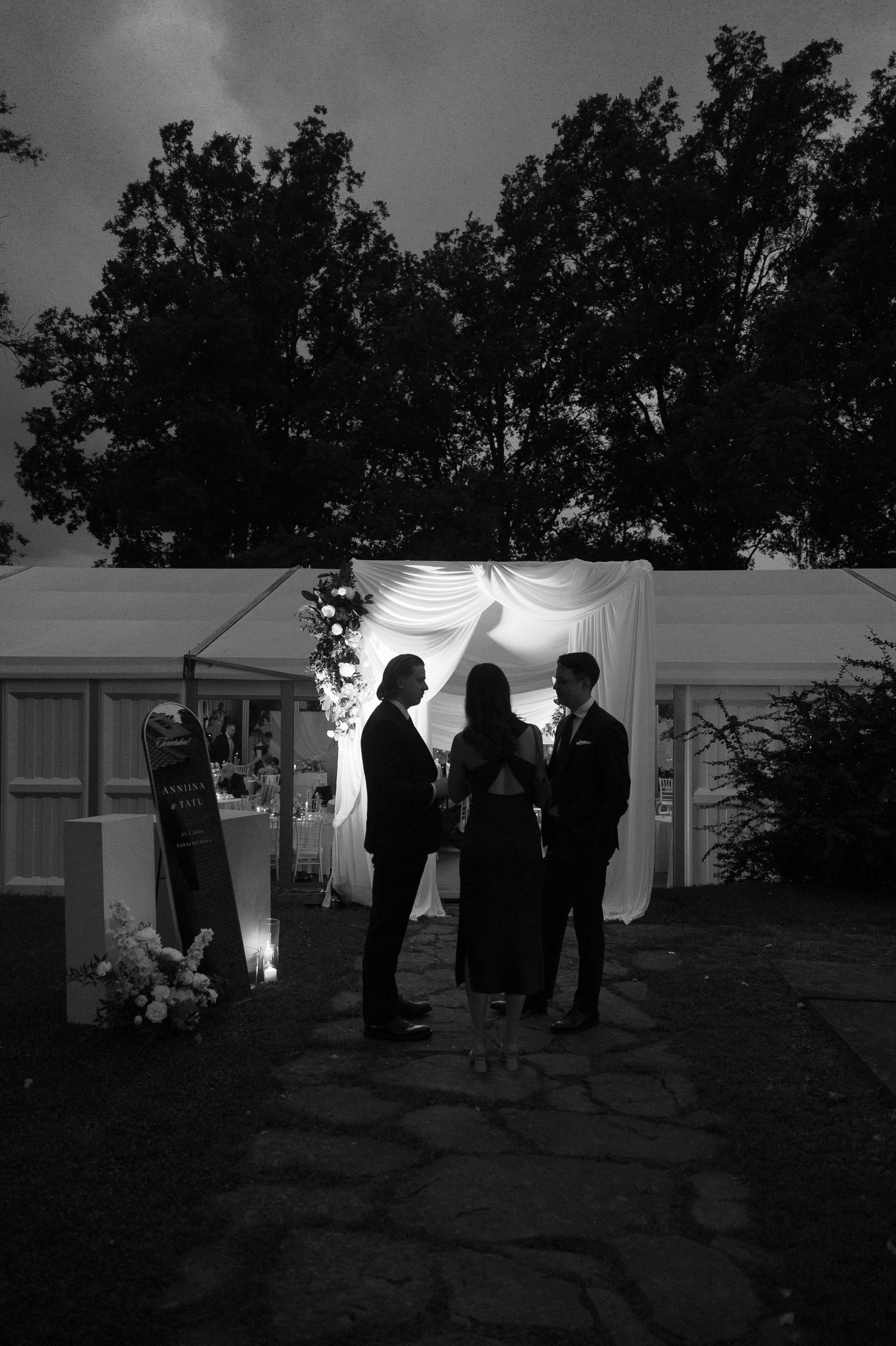 Black and white photo of three silhouetted figures standing outside an illuminated wedding tent at dusk.