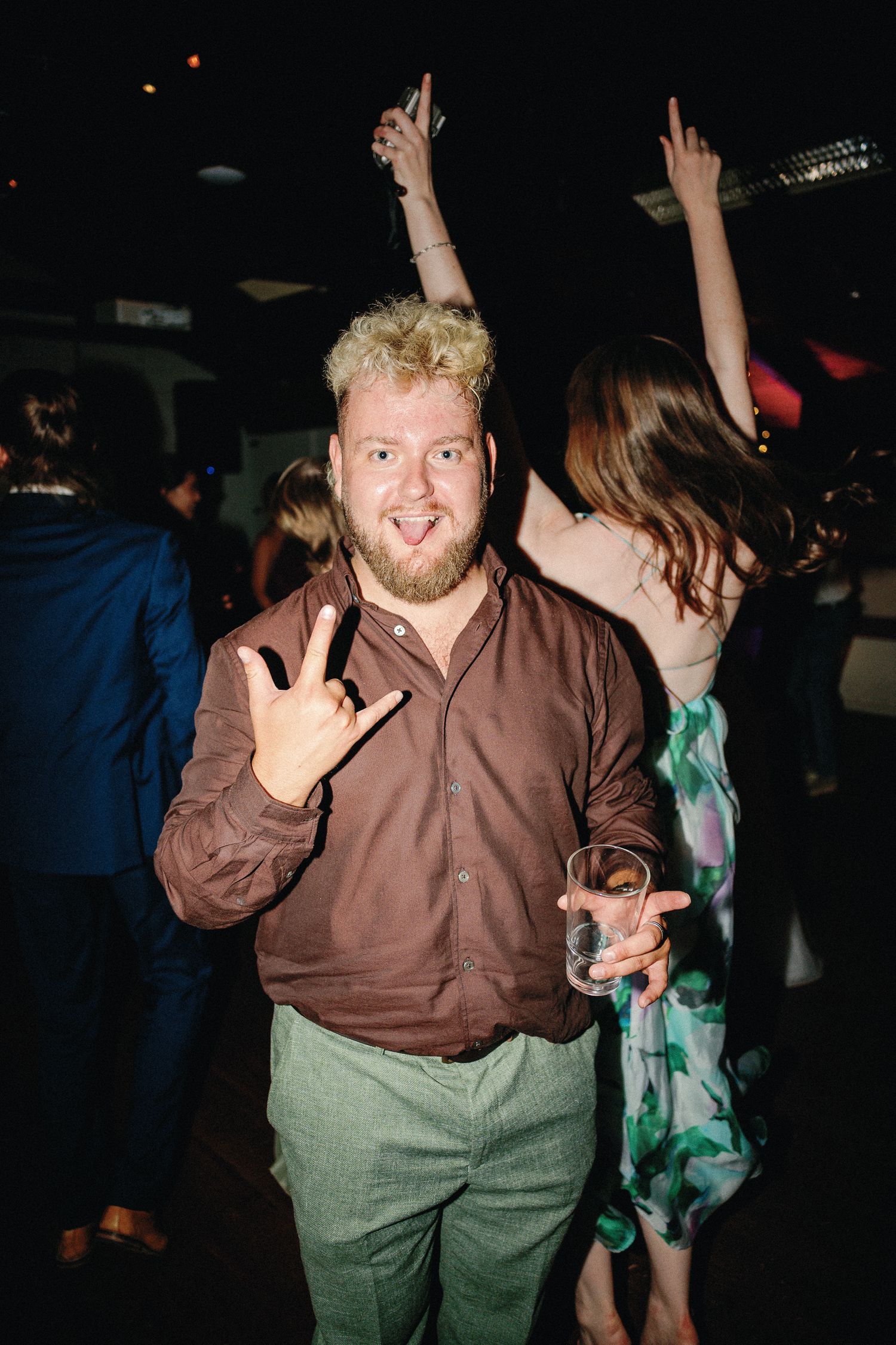 Blond man in brown shirt flashing rock sign at a lively dance party, holding a drink glass.