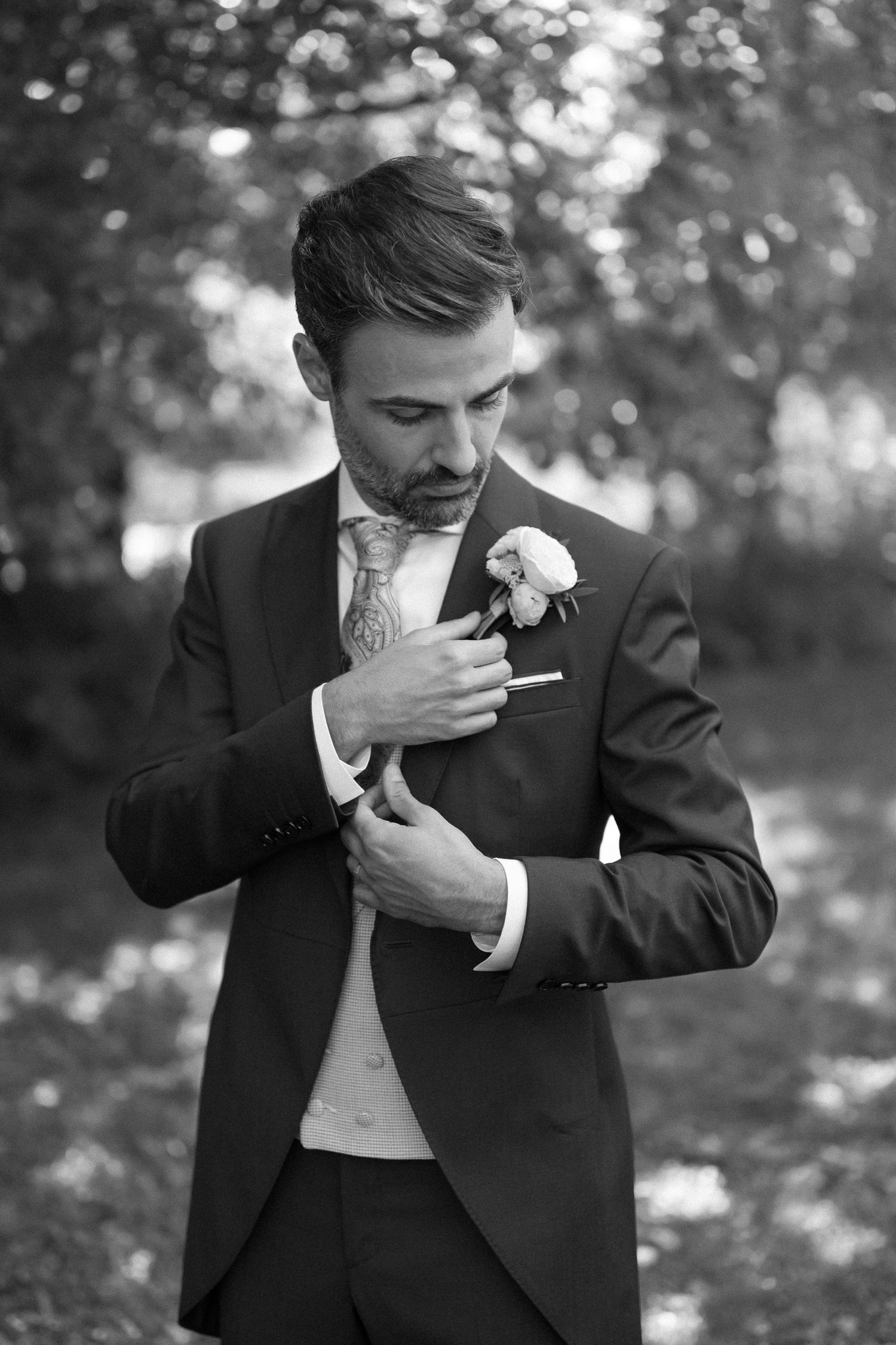 Black and white photo of a groom in a suit adjusting his boutonniere outdoors.