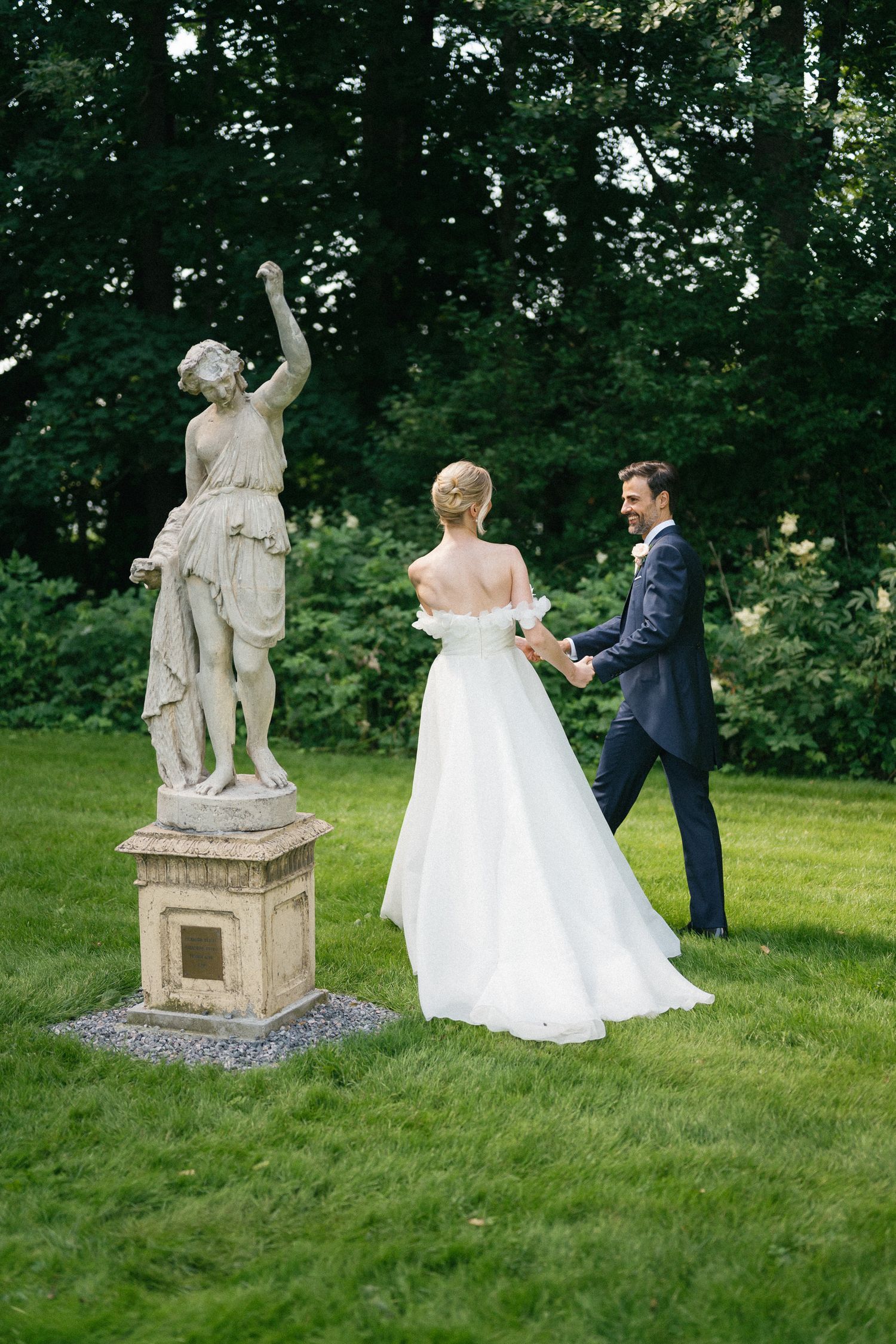 Bride and groom holding hands on lush green lawn beside classical stone statue at elegant outdoor wedding venue.