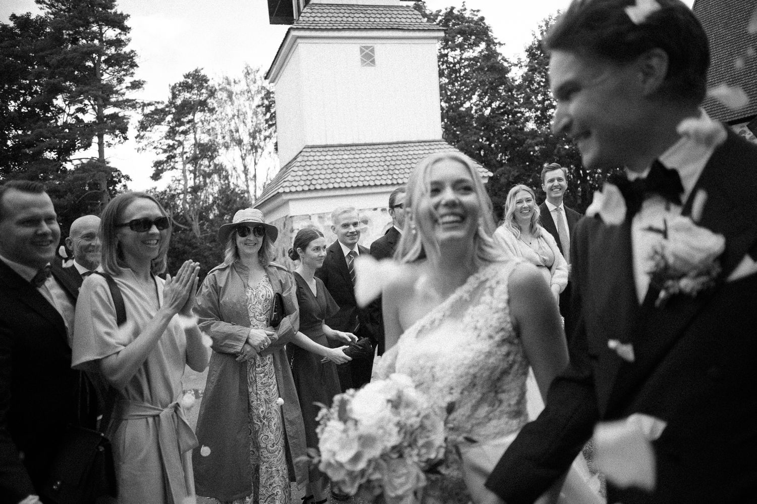 Smiling bride holding a bouquet laughs beside the groom as wedding guests applaud outside a white church in a black and white photo.