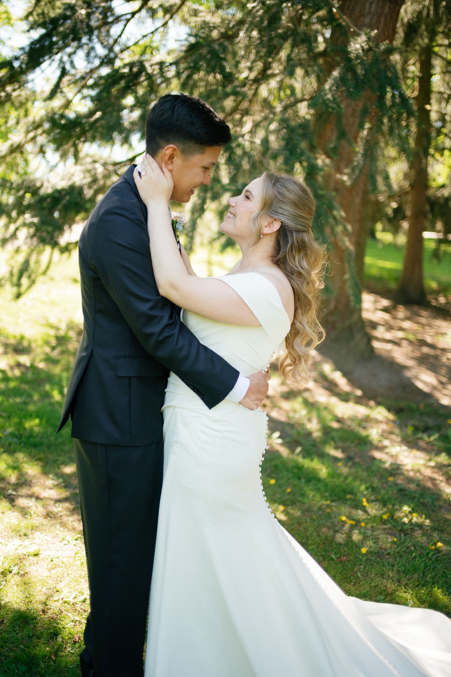 Bride in white gown and groom in black suit embrace romantically outdoors under green trees on their wedding day.