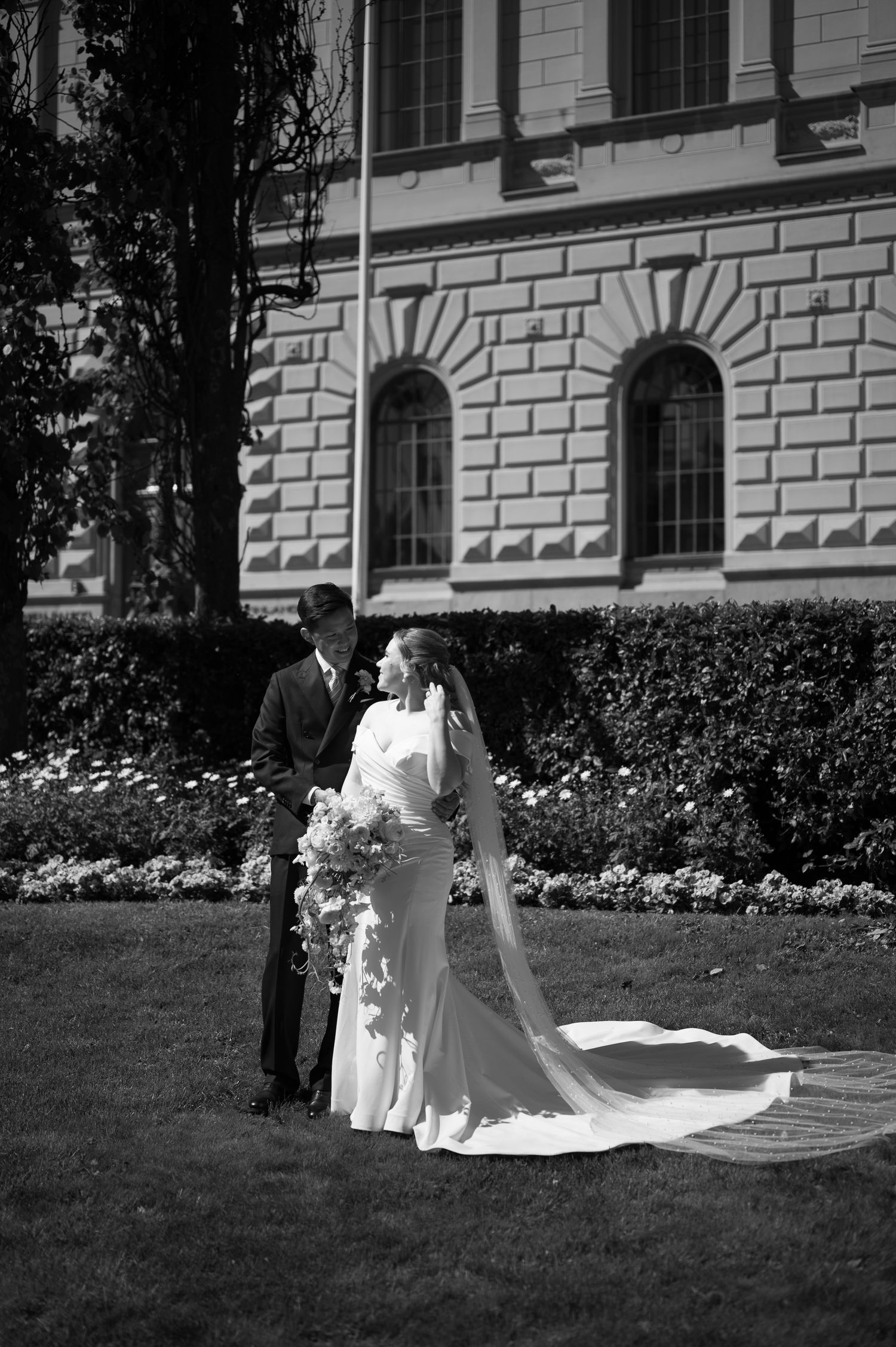 Black and white wedding photo of bride in flowing gown and veil holding bouquet with groom outside historic building.