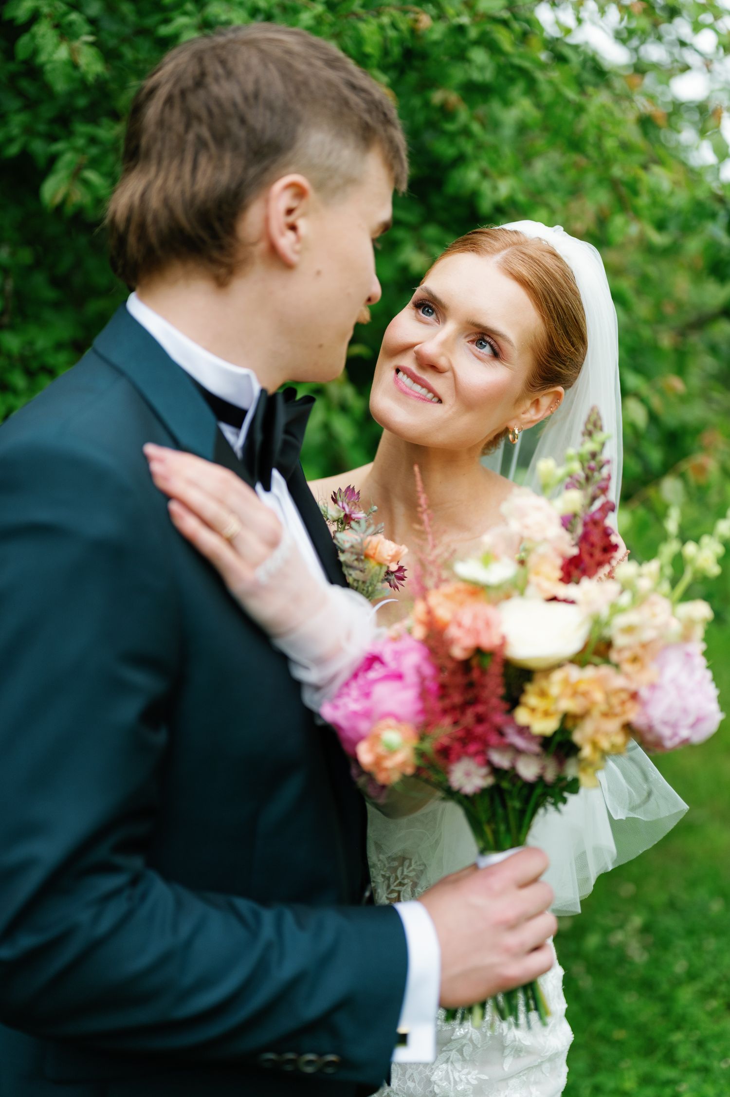 A romantic wedding couple shares an intimate moment outdoors with a colorful bouquet of pink and purple flowers.