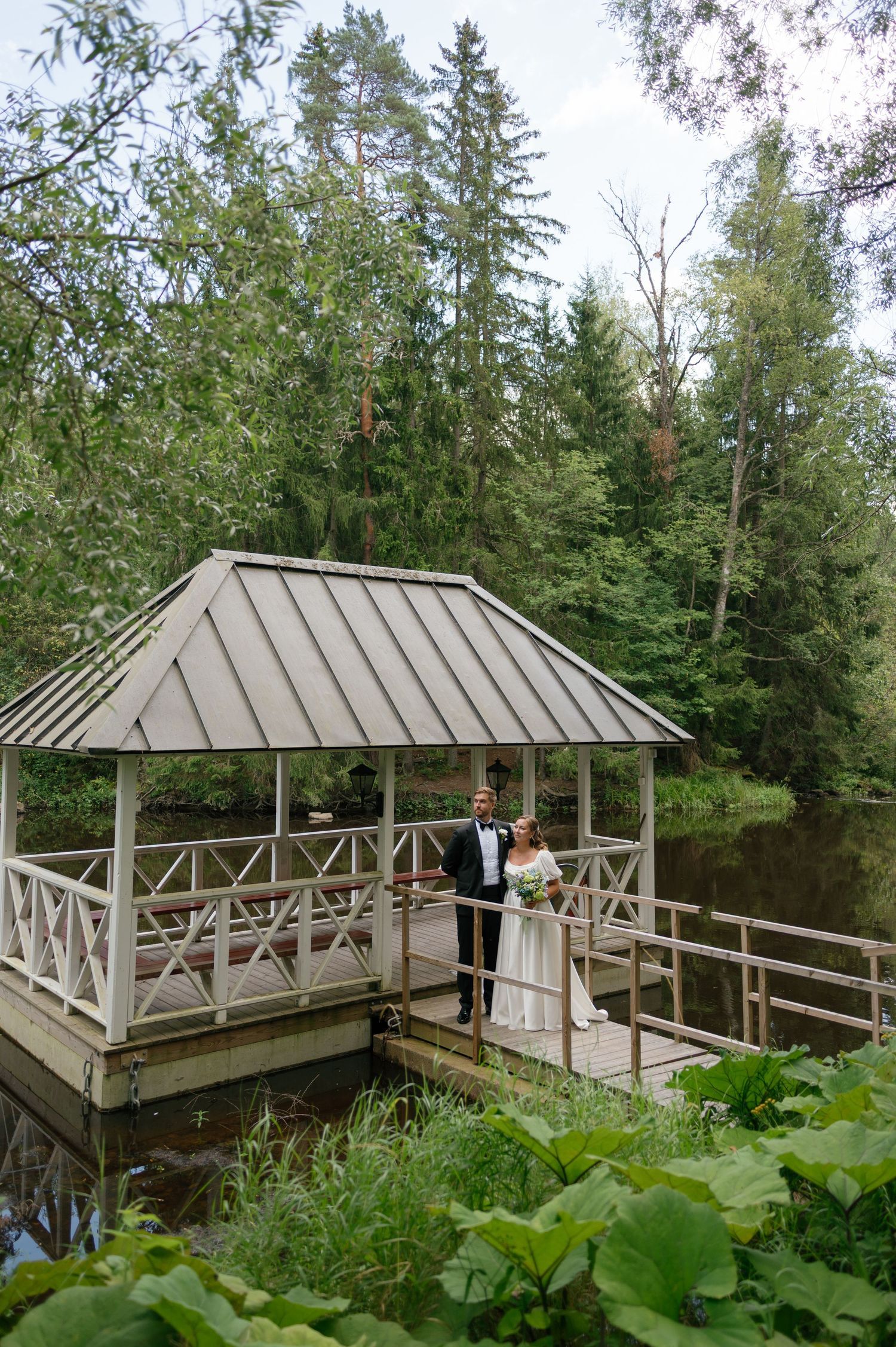 A wedding couple poses in an elegant gazebo overlooking a tranquil pond surrounded by evergreens.