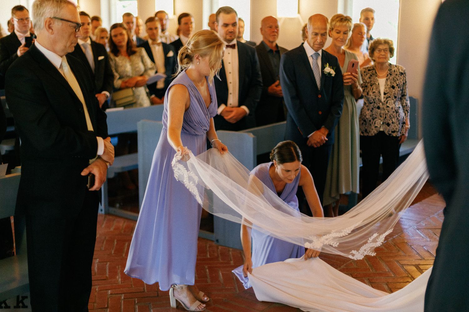 Attendants in lavender dresses adjust the bride's train during the wedding ceremony.