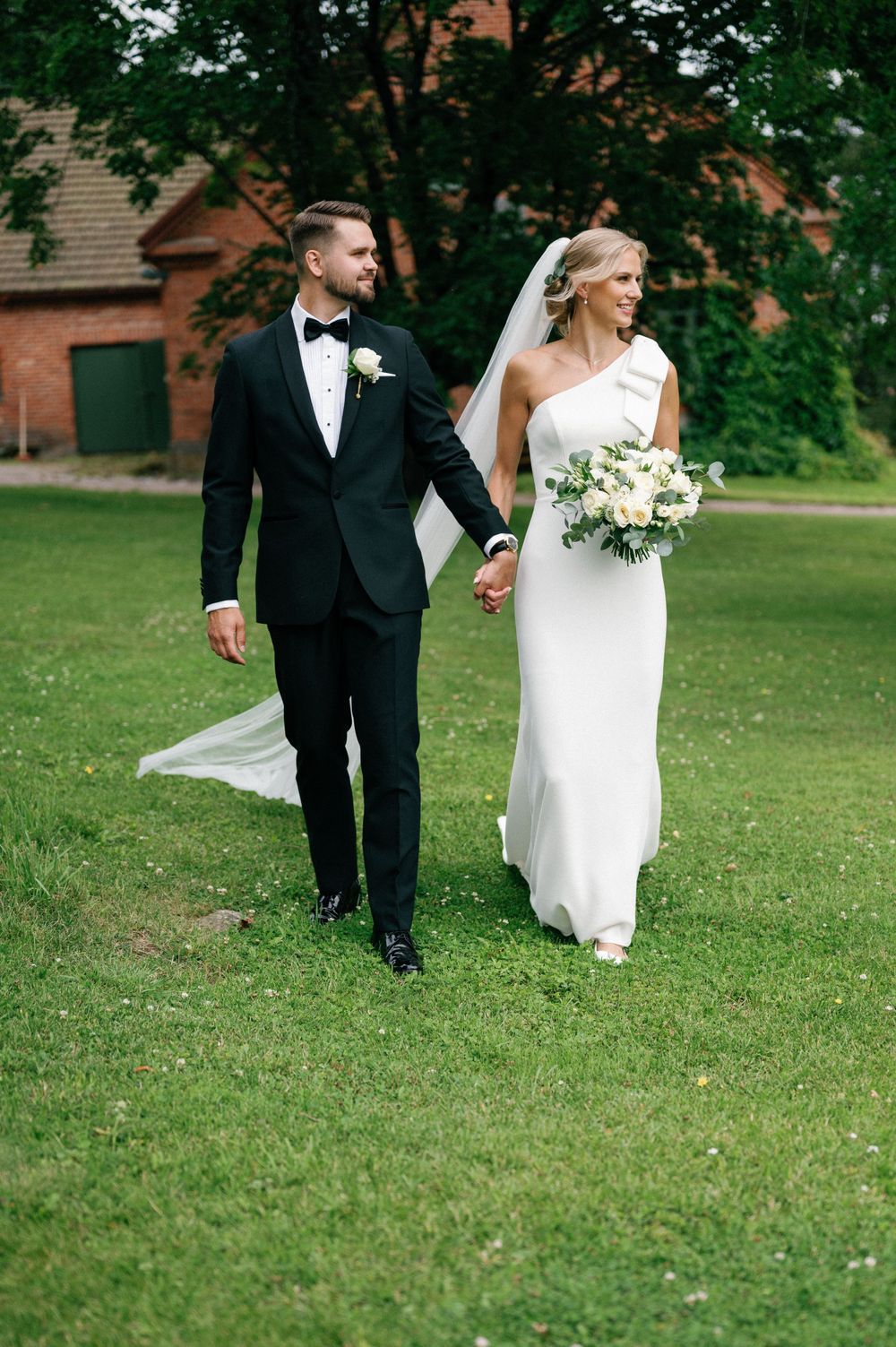 A couple in wedding attire strolls together on manicured grass with trees in the background.