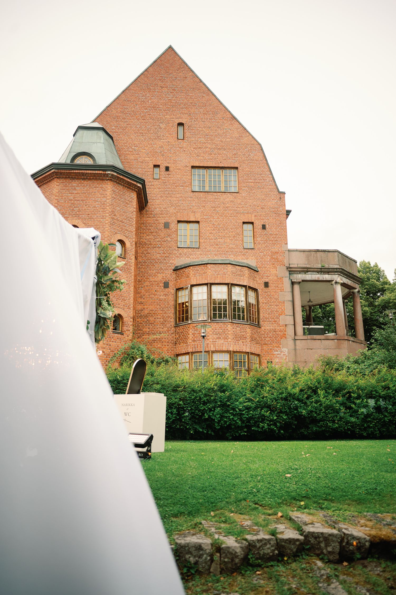 Historic red brick building with arched windows and pointed gable roof surrounded by lush green lawn and shrubs.