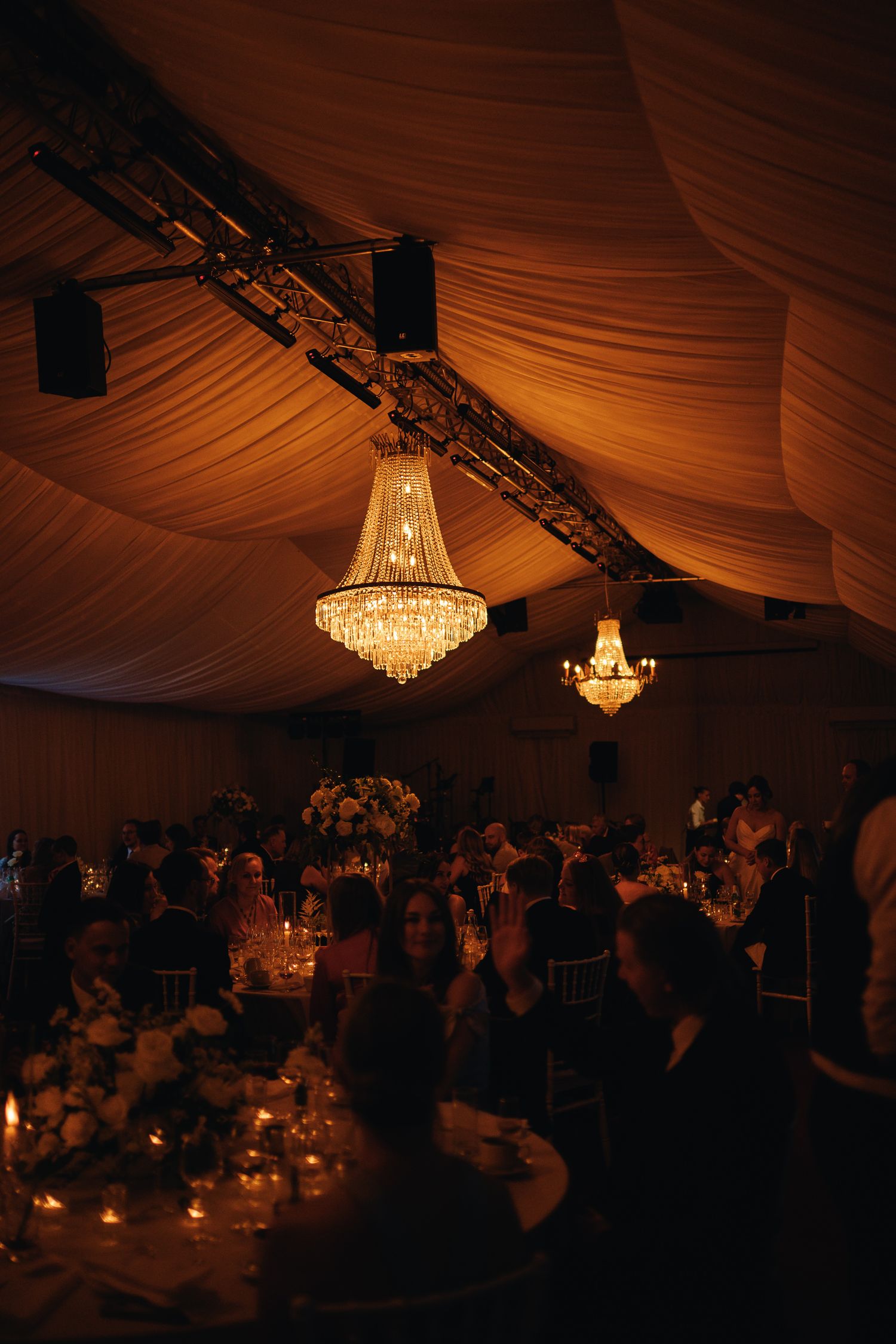 Elegant wedding reception in a draped tent with crystal chandeliers, candlelit tables, and floral centerpieces.