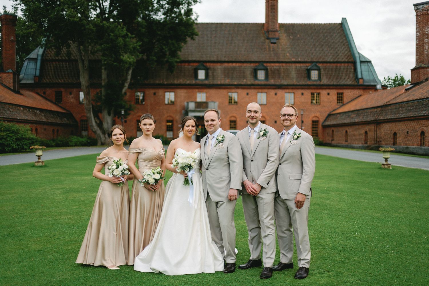 Wedding party posing outdoors in front of a red brick venue, bridesmaids in champagne gowns and groomsmen in grey suits.