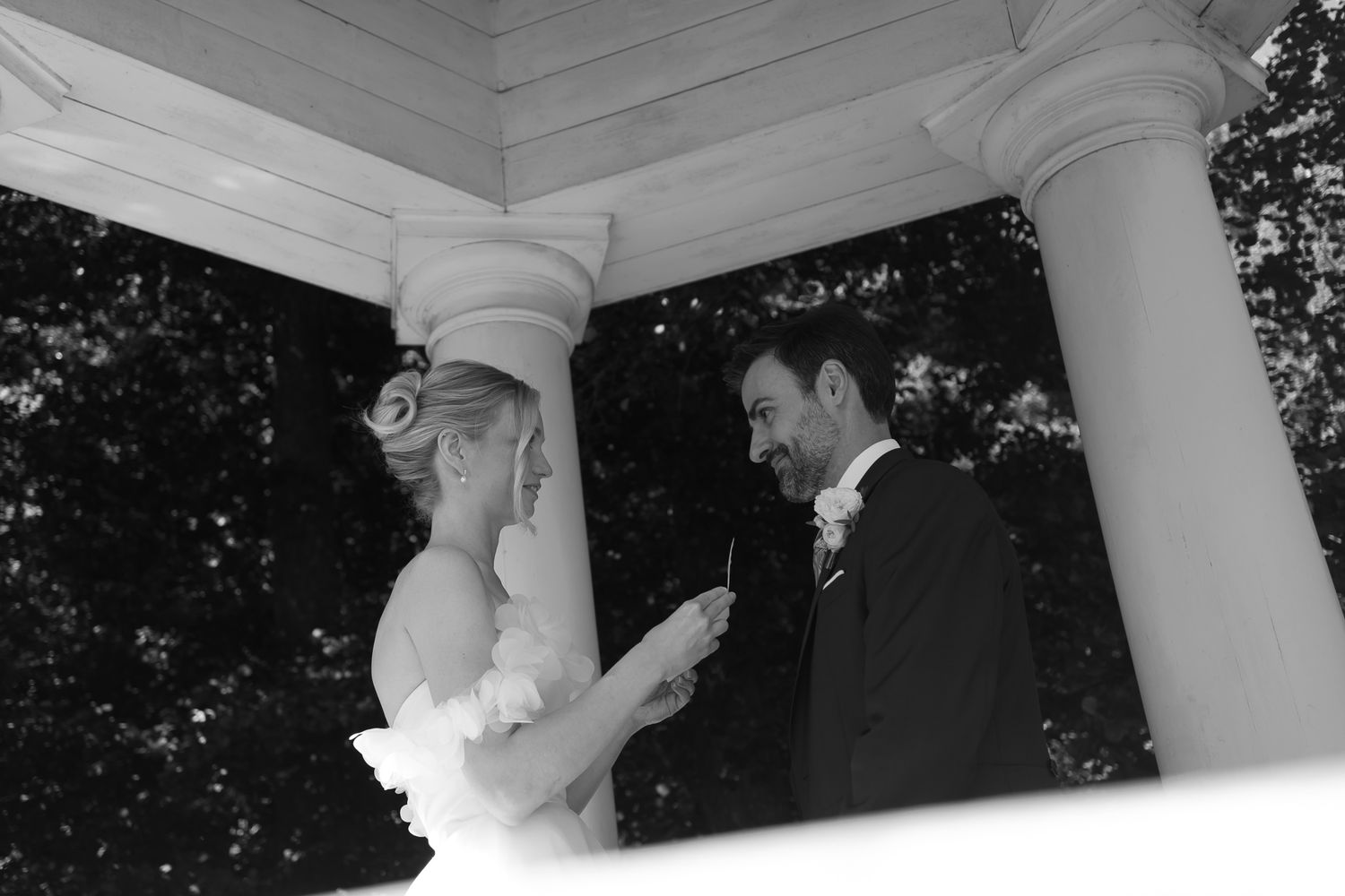 Black and white wedding photo of bride and groom exchanging vows in an elegant white gazebo surrounded by trees.