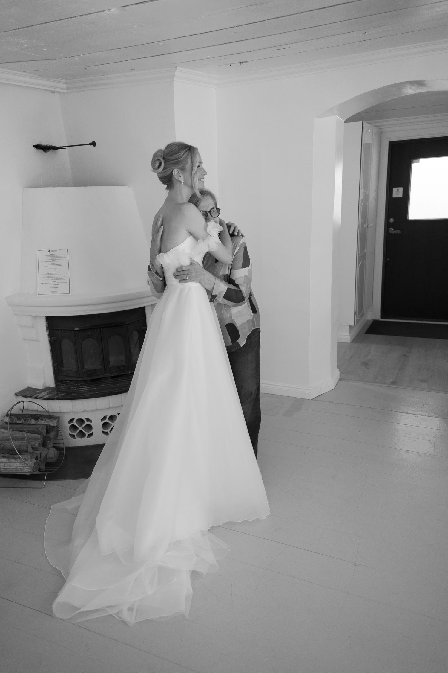 Black and white wedding photo of bride in elegant gown embracing partner near fireplace indoors.