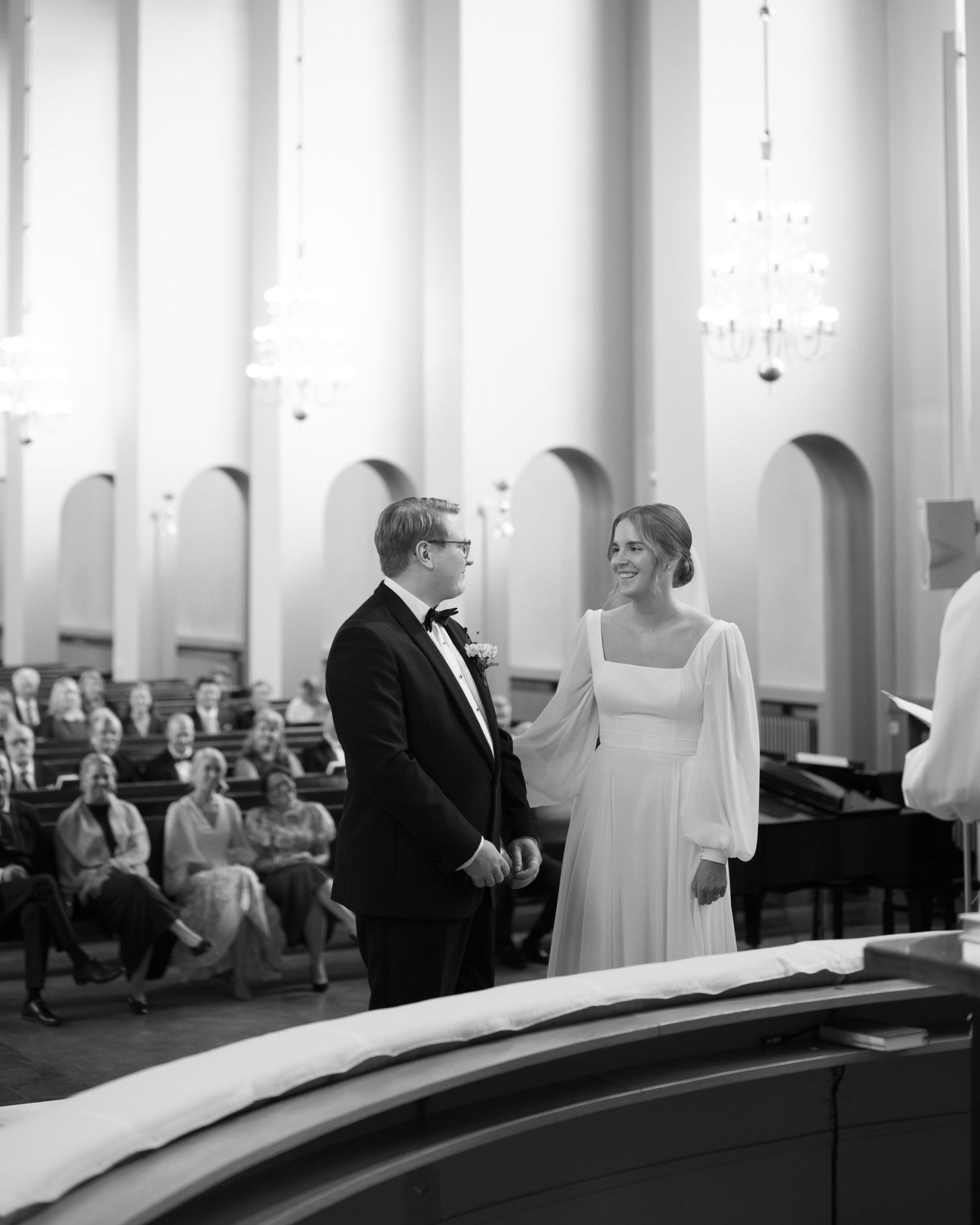 A black and white wedding ceremony scene with guests seated in an elegant room with arched architectural details.