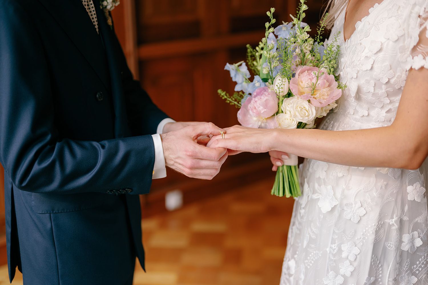 Close-up of hands exchanging rings with a pastel bouquet during a wedding ceremony.