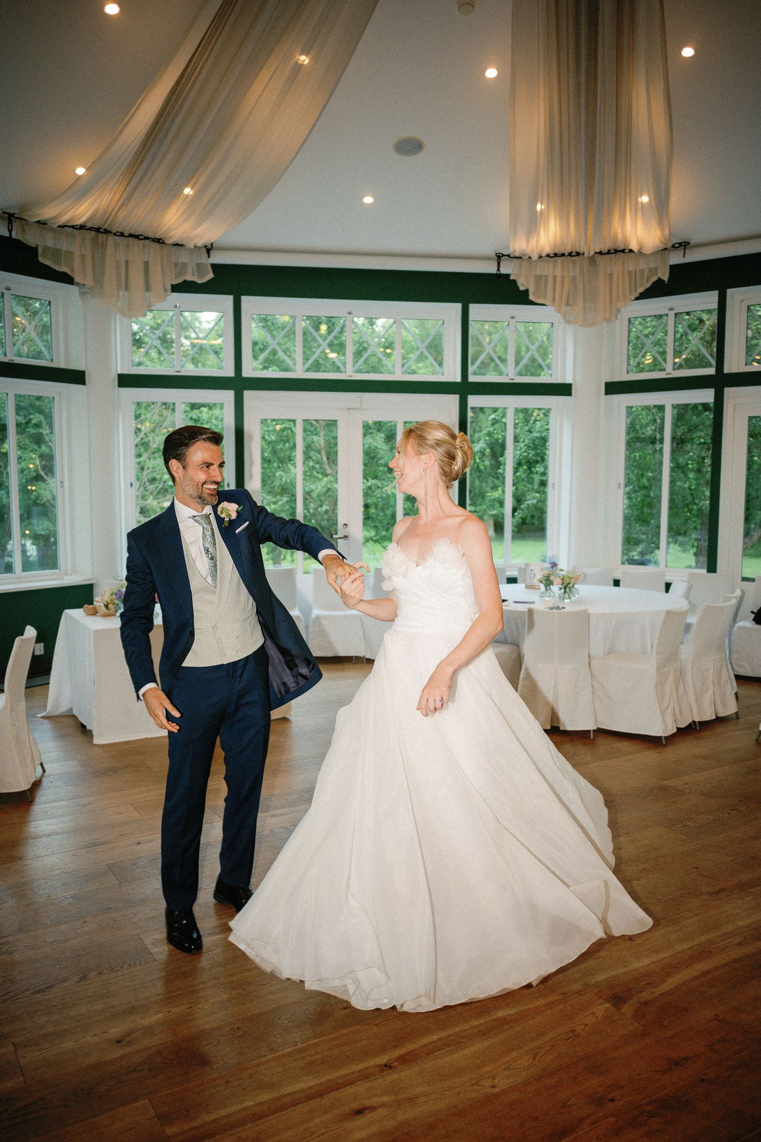 Bride and groom share their first dance in an elegant wedding venue with large windows and fairy lights.