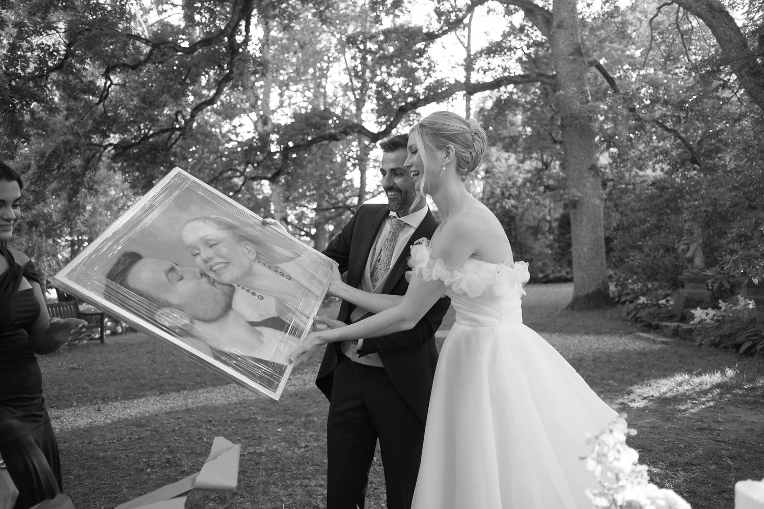 Black and white wedding photo of bride and groom holding a framed baby portrait outdoors.