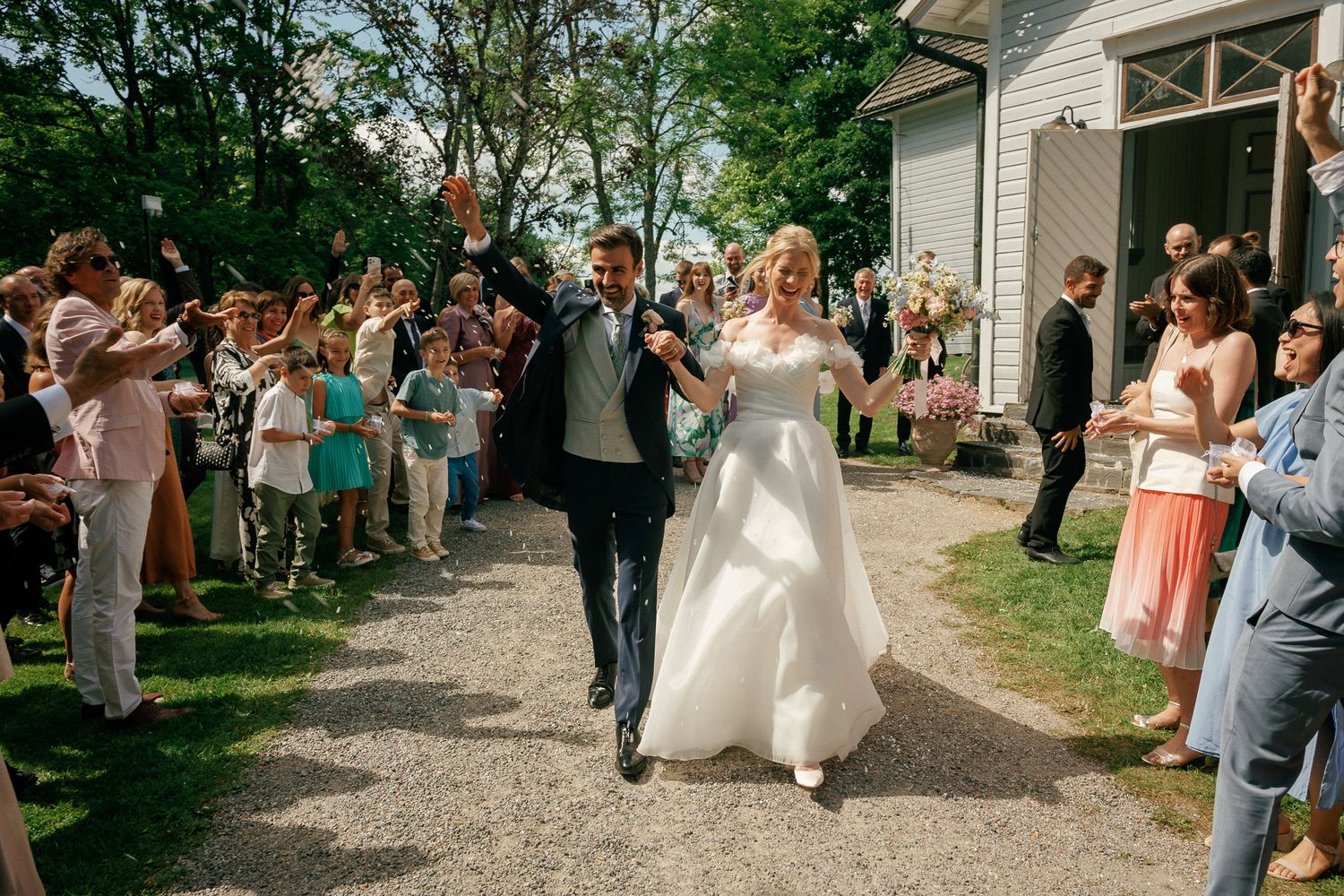 Bride and groom walk joyfully through cheering guests outdoors at a sunny wedding reception celebration.