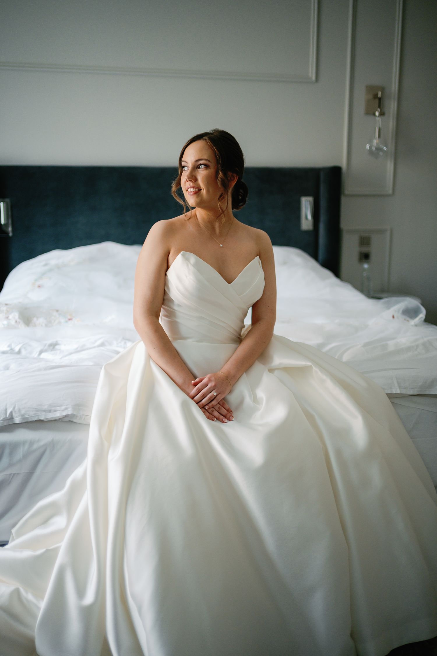 Bride in strapless white ballgown sitting on hotel bed, smiling softly, with elegant updo hairstyle.