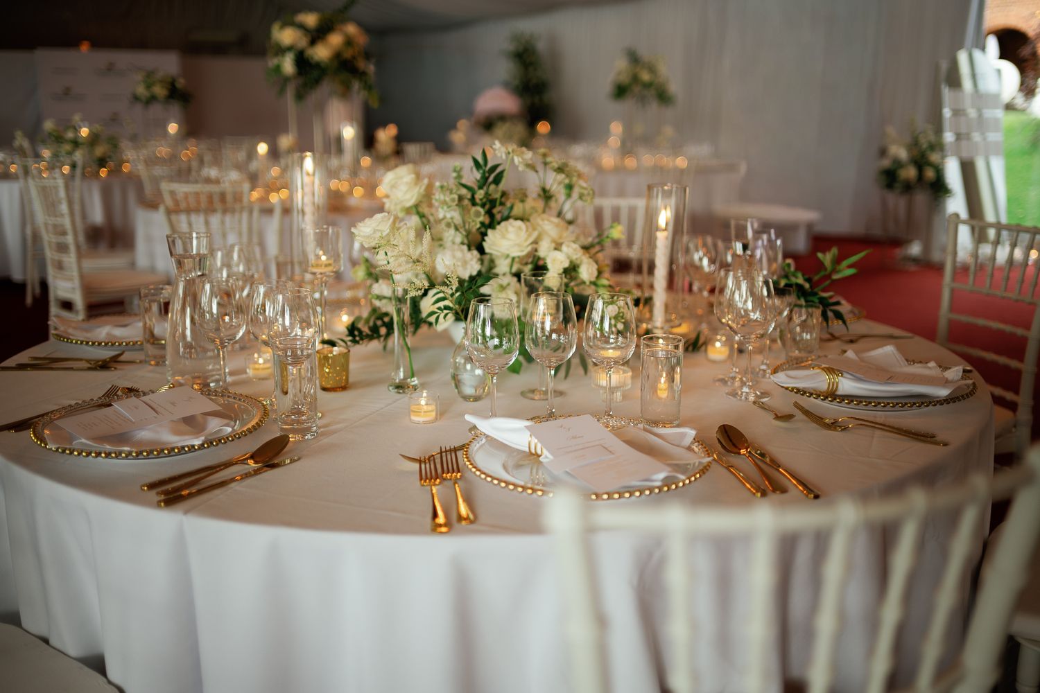 Elegant wedding reception table with white floral centerpiece, gold cutlery, crystal glasses, and candles on ivory linen.