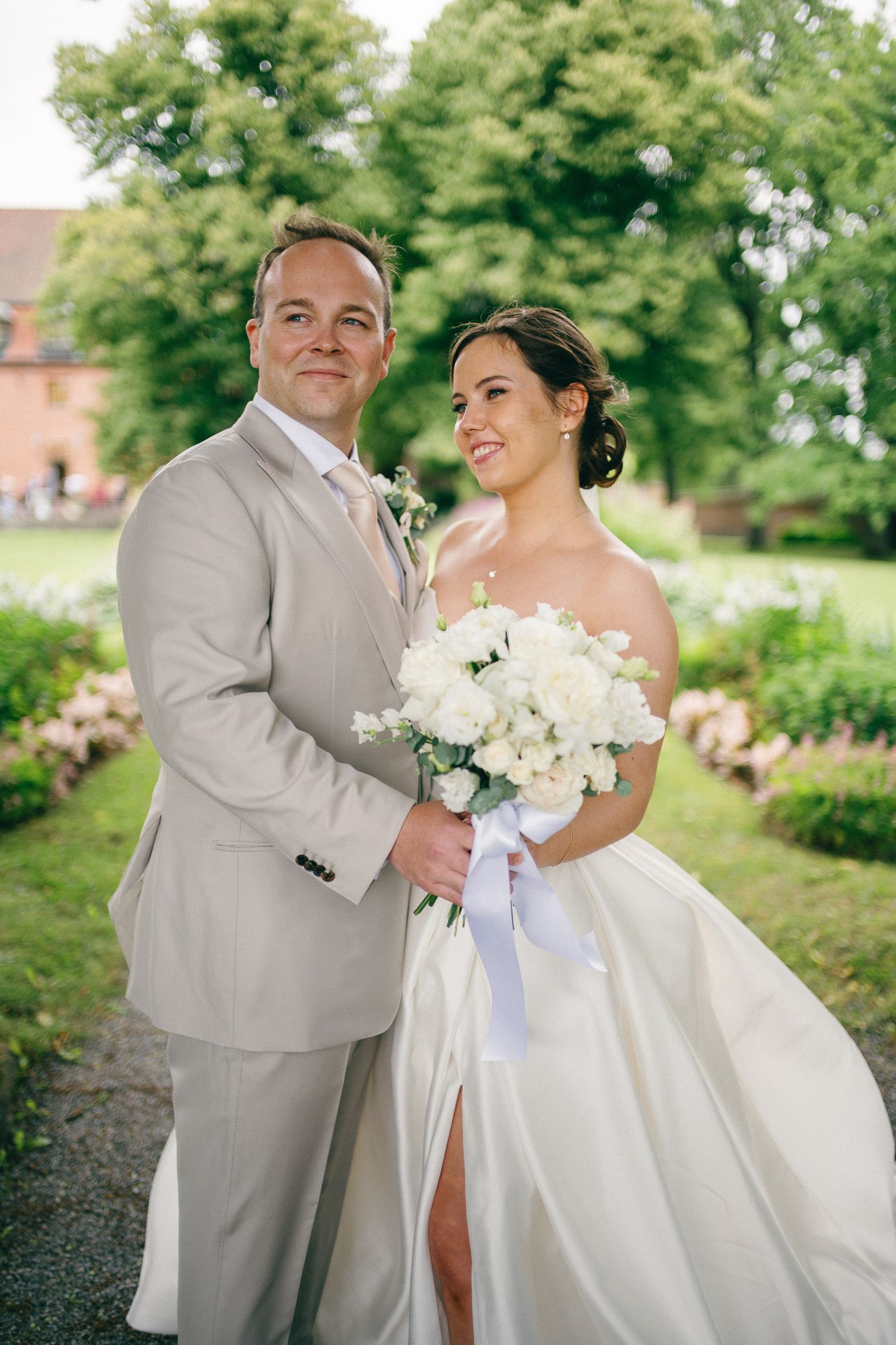 Bride and groom posing outdoors in lush garden, bride holding white bouquet in strapless gown, groom in beige suit.