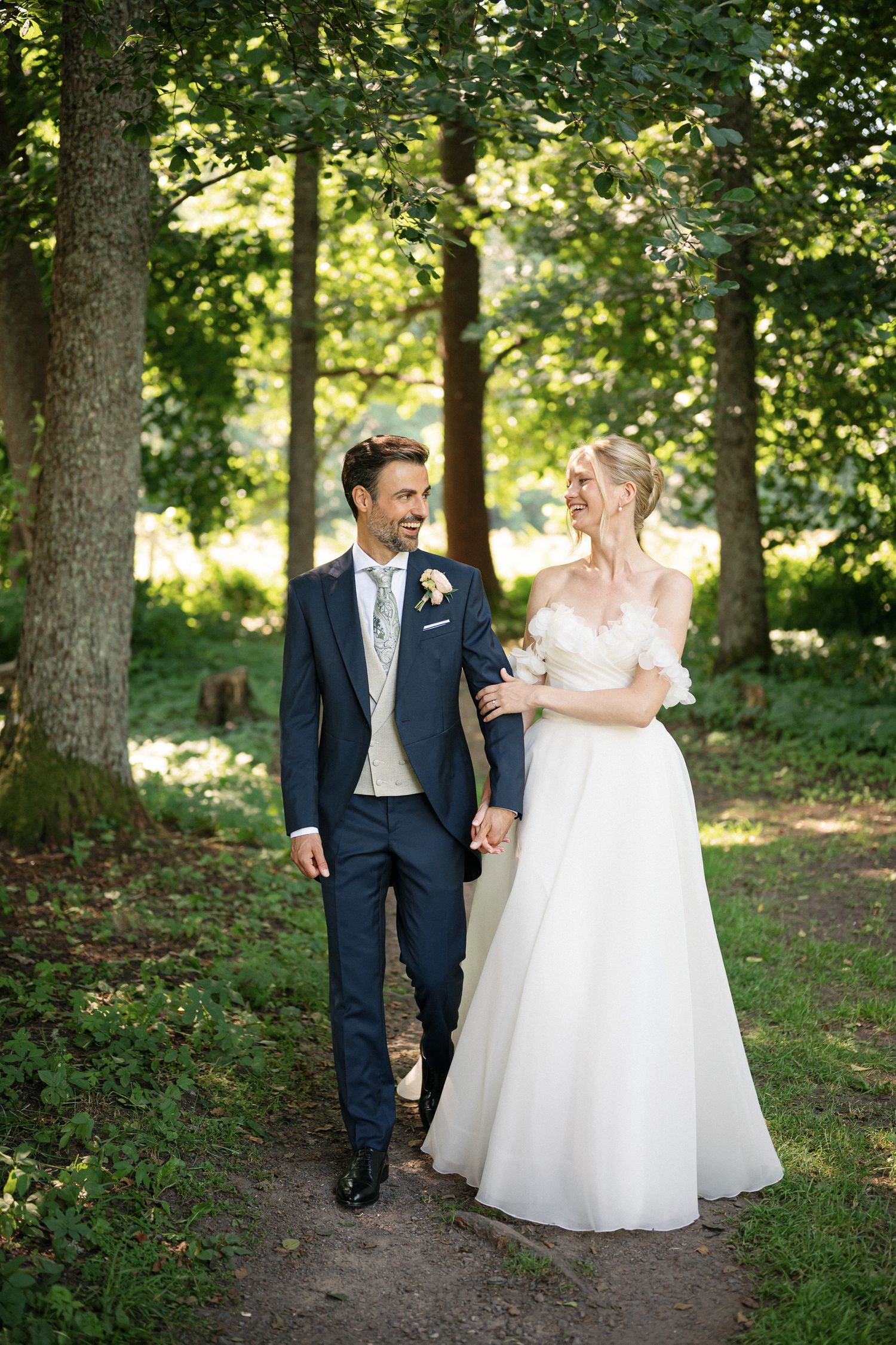 Bride and groom walking hand in hand through a lush green forest path on their wedding day.