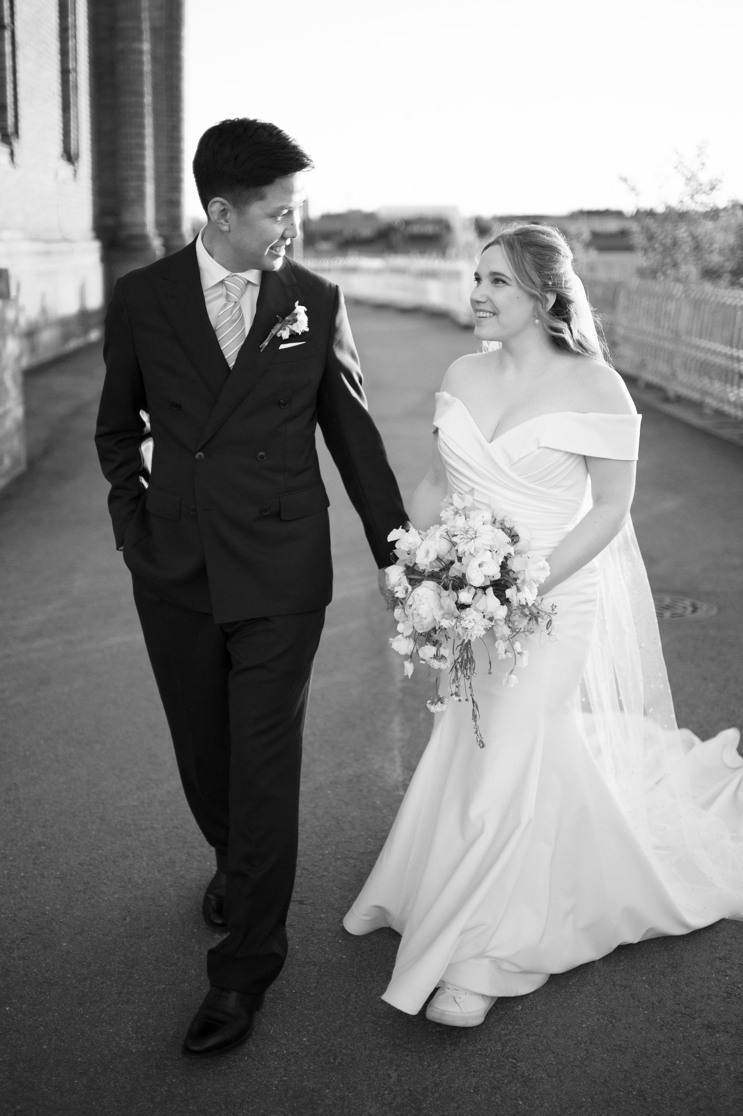 Black and white photo of a smiling bride and groom walking together on their wedding day, bride holding a floral bouquet.