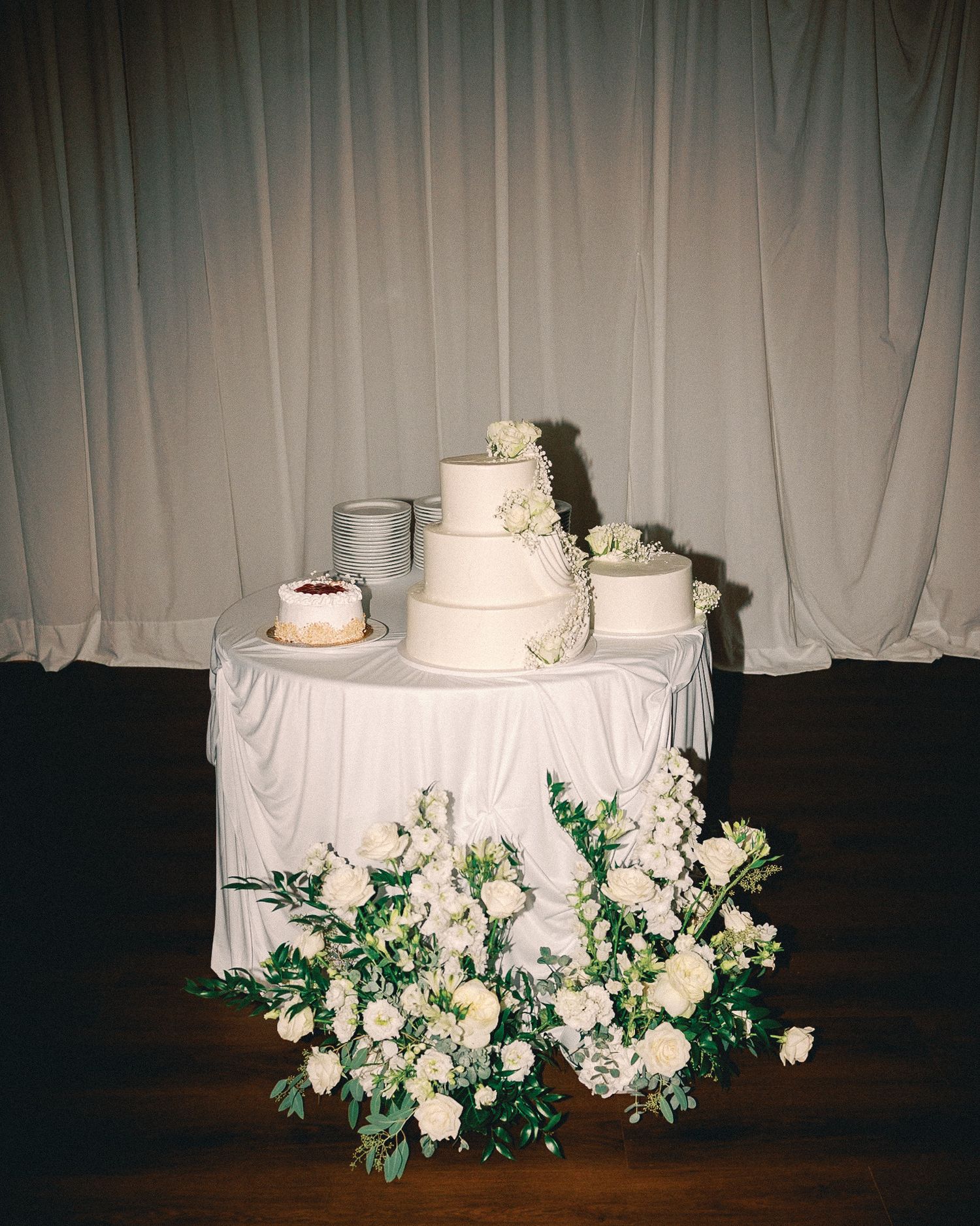 Elegant three-tier white wedding cake with floral decorations and white roses arrangement on a draped table.