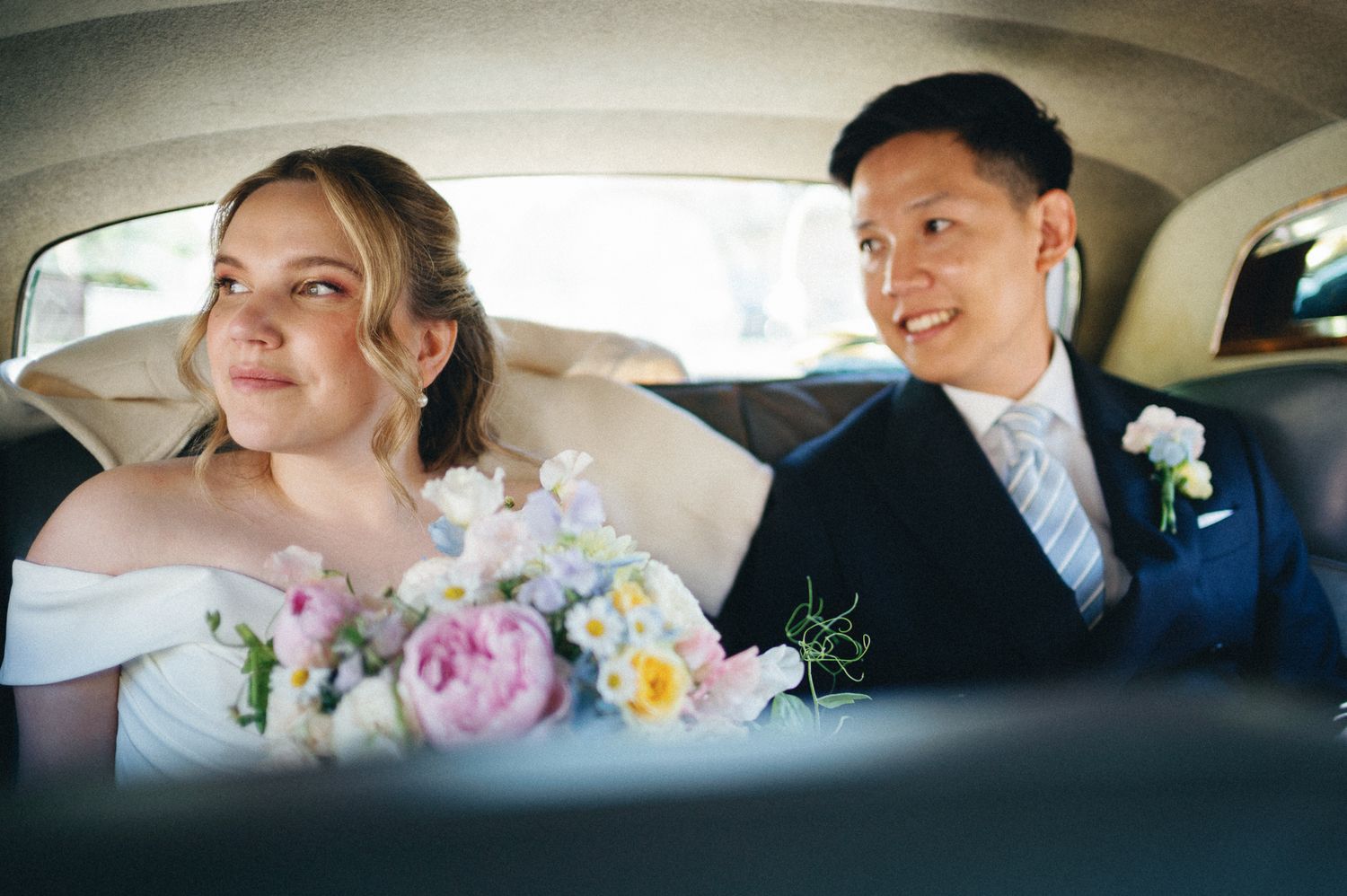 Bride and groom sitting in backseat of wedding car, bride holding pastel floral bouquet, groom in navy suit.