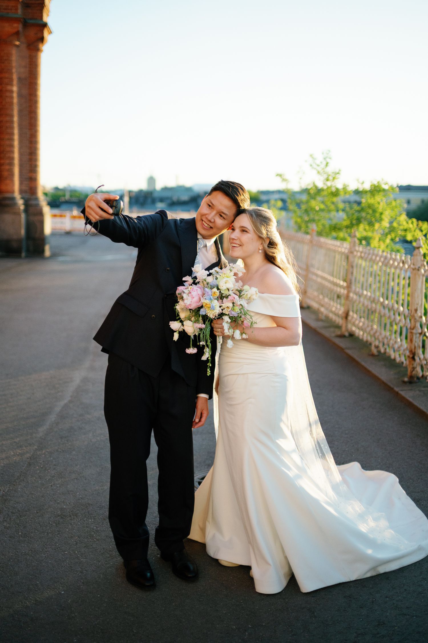 Bride and groom taking a selfie outdoors, bride in off-shoulder gown holding floral bouquet, groom in black tuxedo.