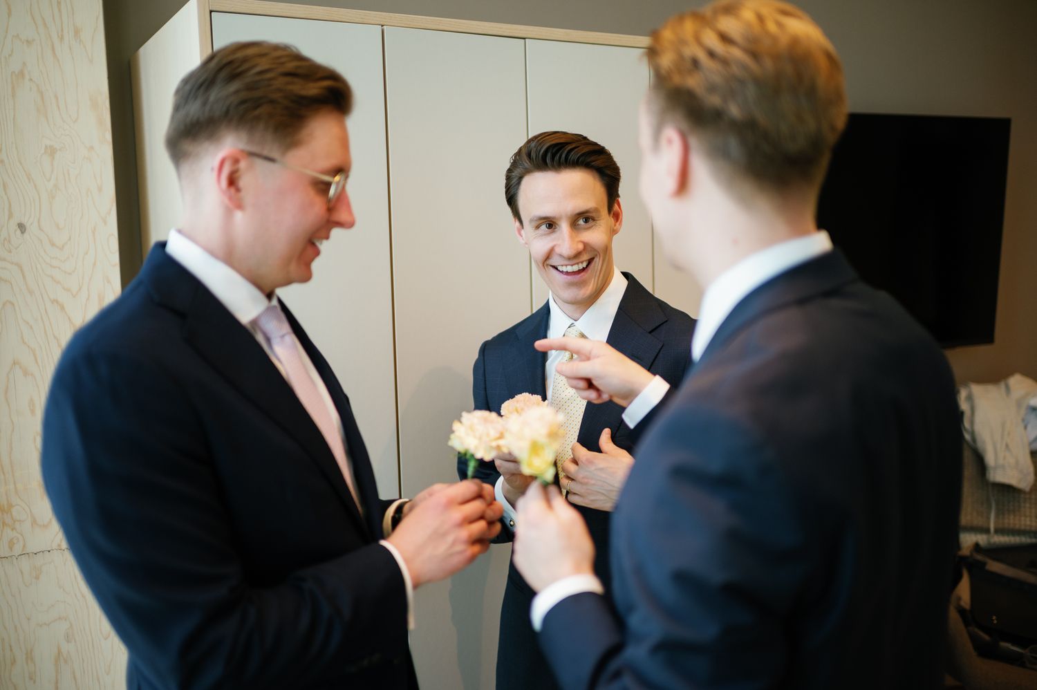 Three groomsmen in navy suits arranging boutonnieres before a wedding ceremony.