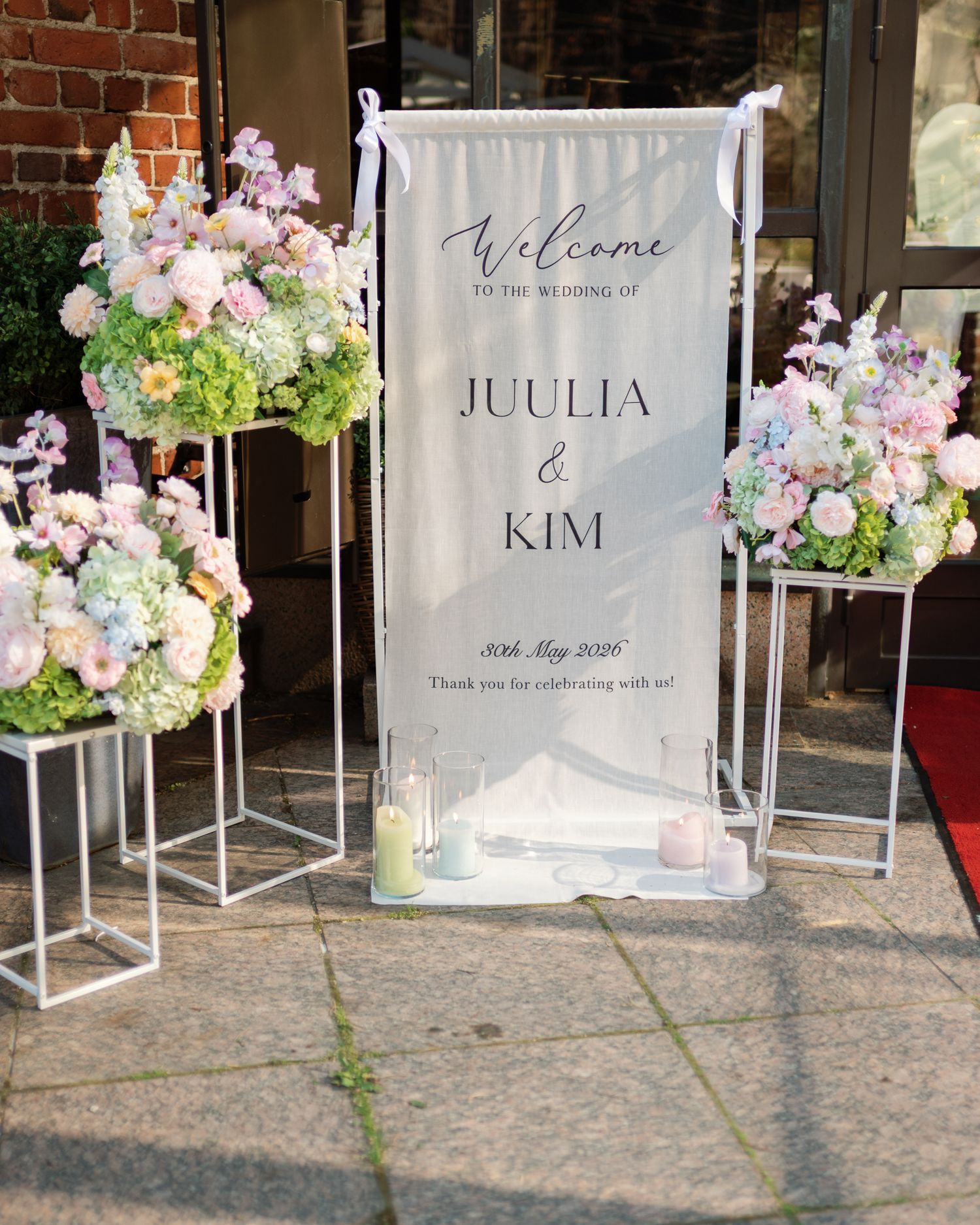 Wedding welcome sign for Juulia and Kim flanked by pastel floral arrangements on white geometric stands outdoors.