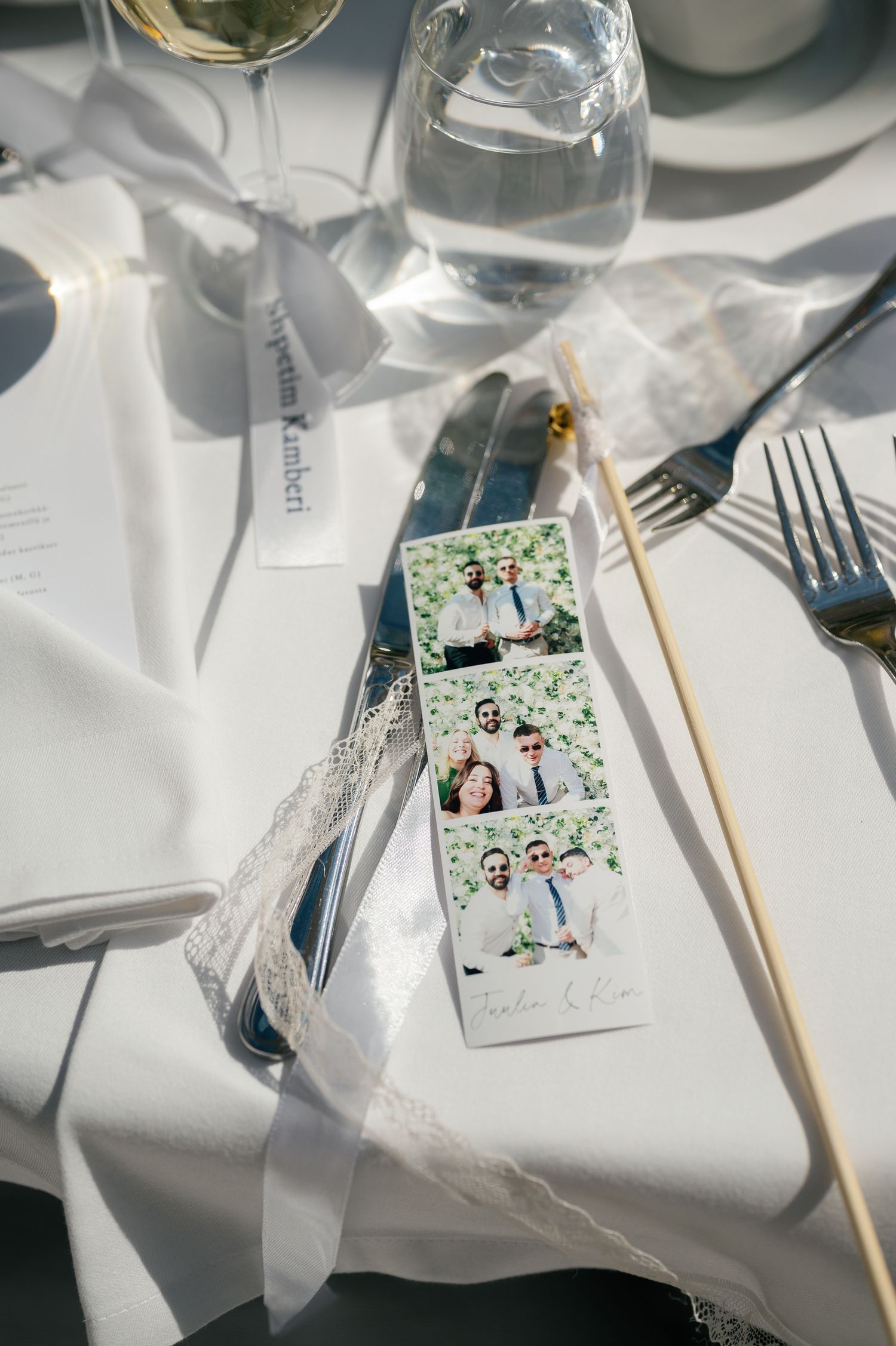 Elegant wedding table setting with photo booth strip, lace ribbon, gold skewer, cutlery, and glassware on white linen.